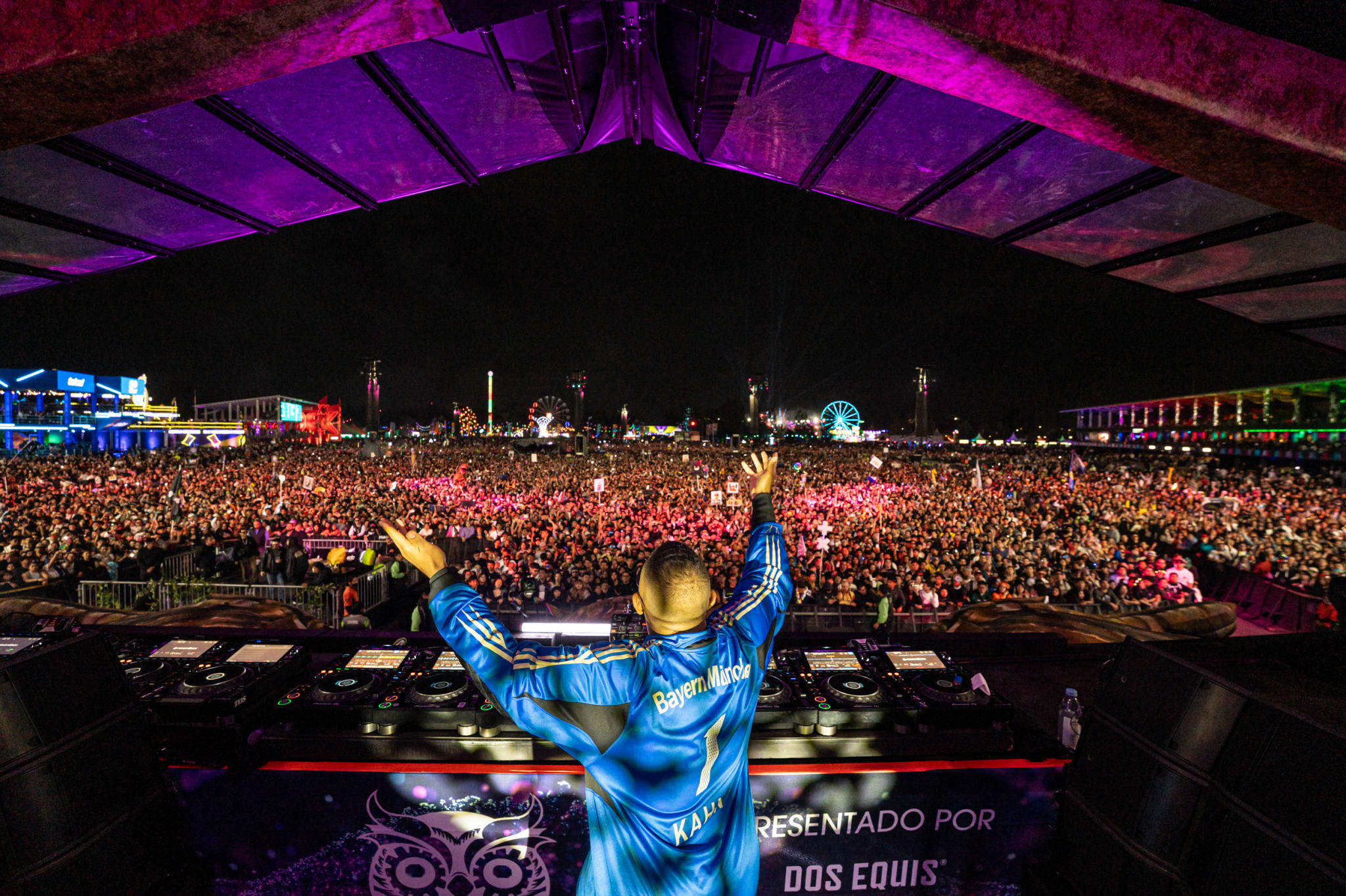 Gordo performing on stage at a large outdoor music festival, with a vibrant, colorful crowd and a Ferris wheel in the background.