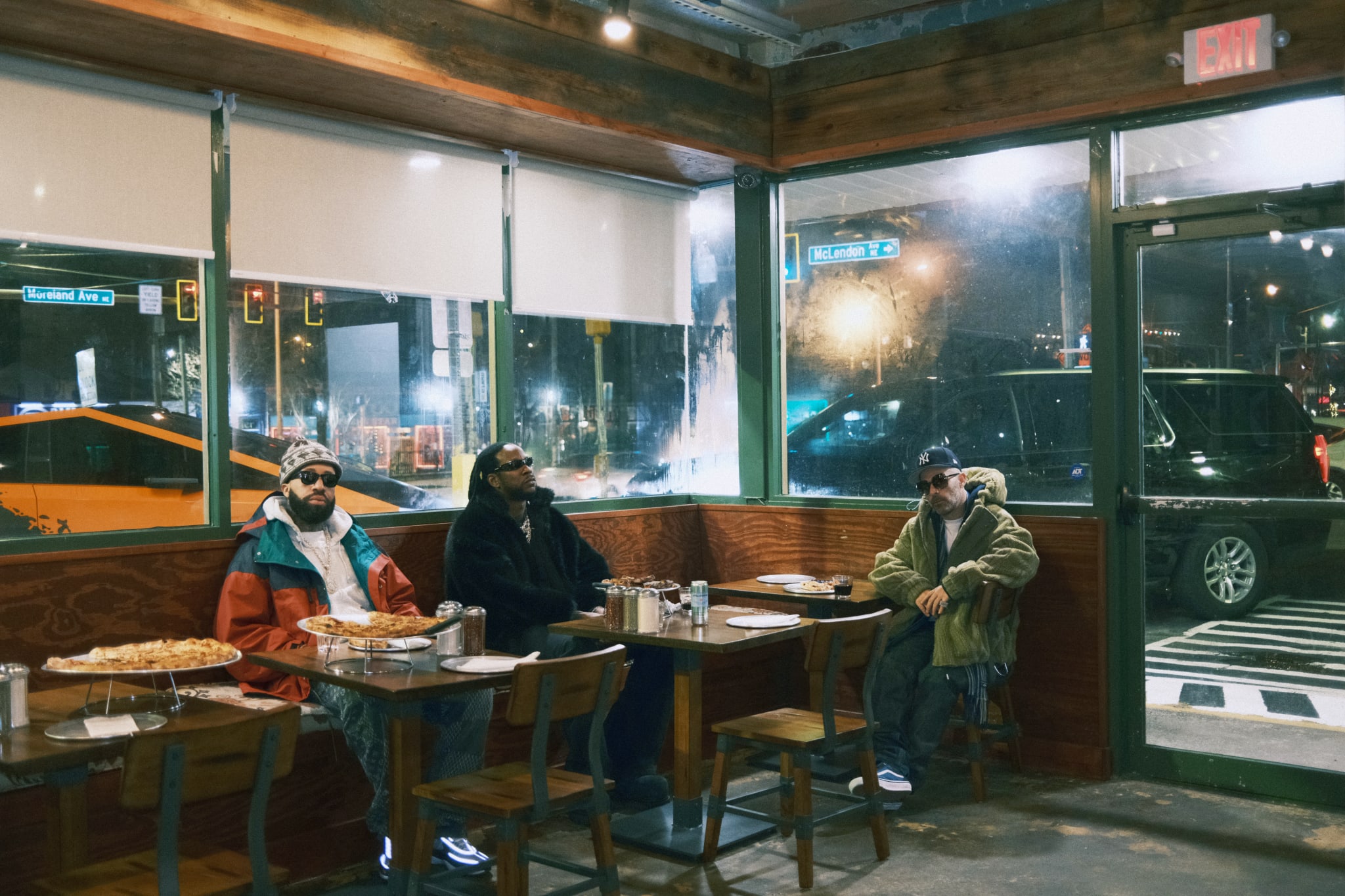 Larry June, 2 Chainz and Alchemist wearing winter clothing and sunglasses sitting at a pizzeria table with pizza and drinks, in a cozy, dimly lit restaurant.