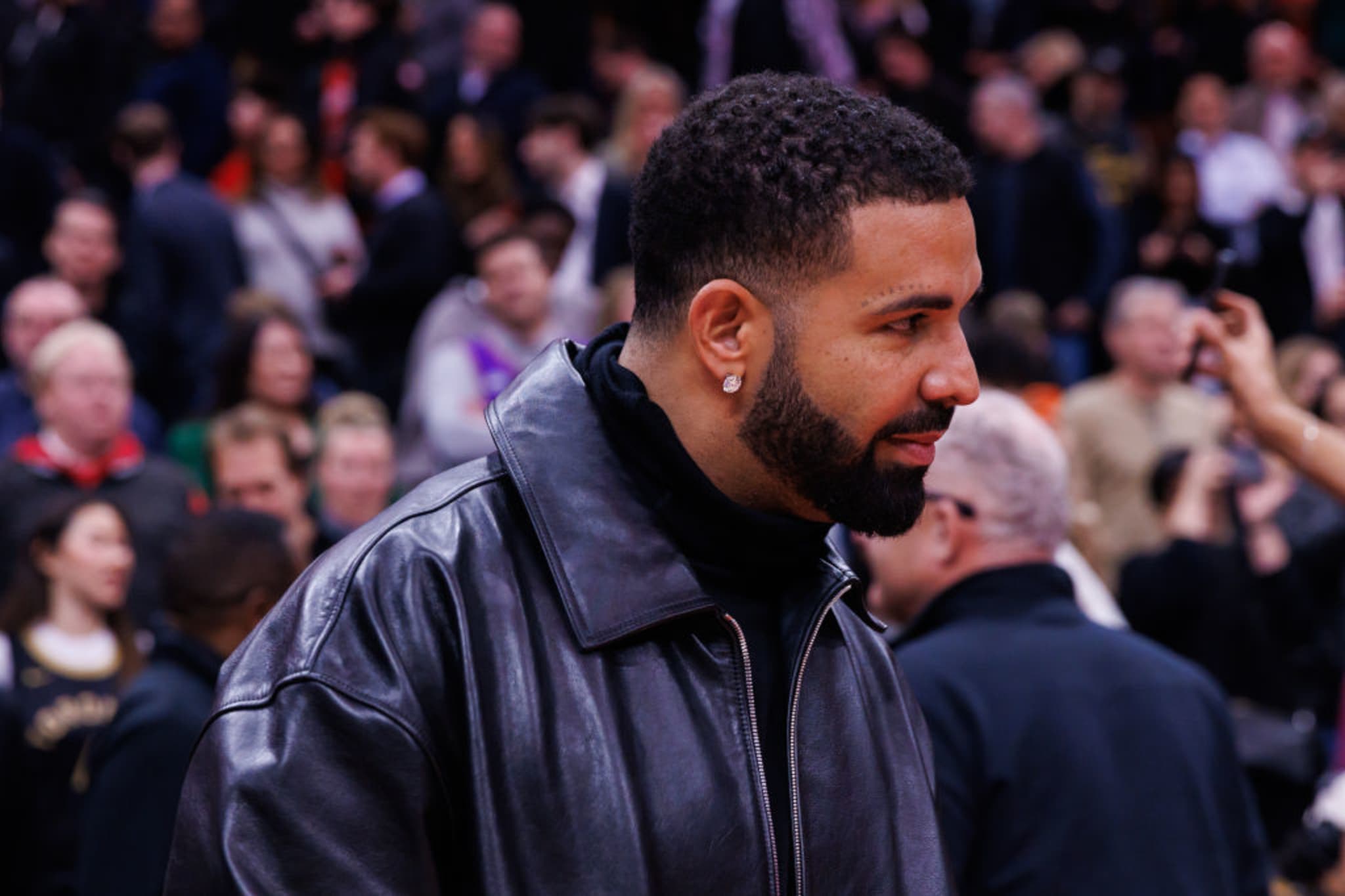 Drake wearing a black leather jacket, standing in a crowded setting, looking to the side.