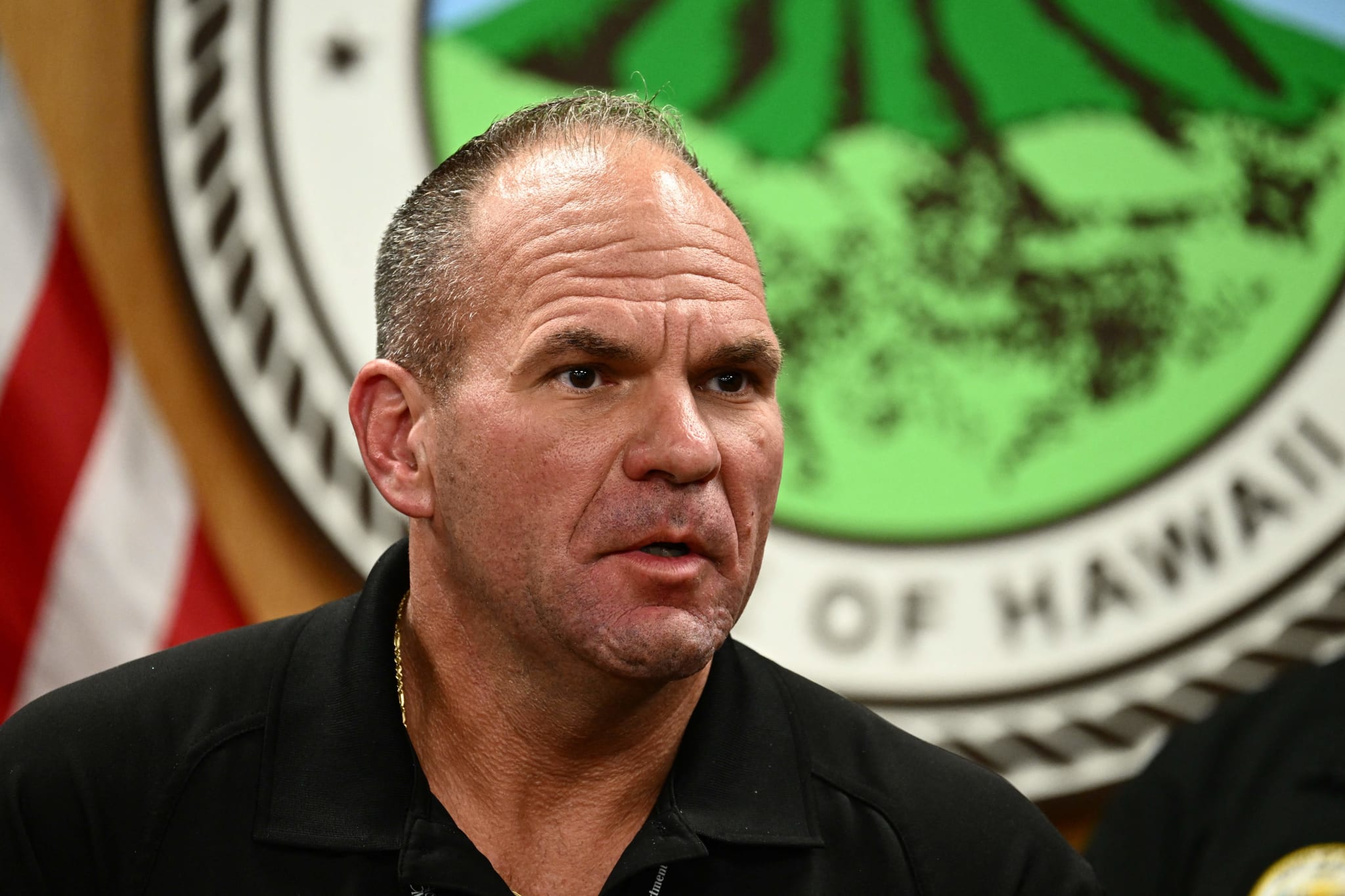 Maui Police Chief John Pelletier speaking in front of a seal with "Hawaii" visible, wearing a black shirt.