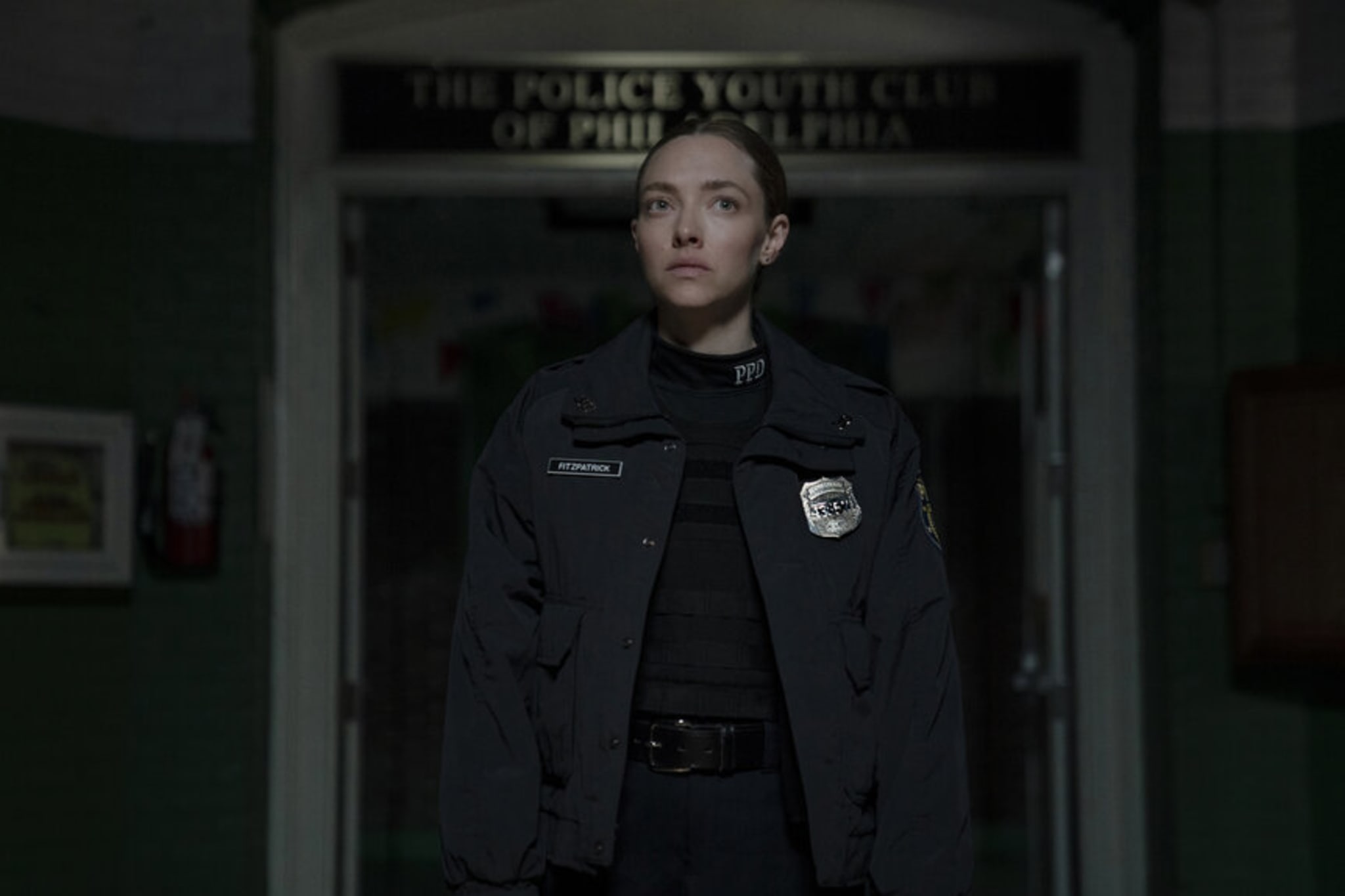 Amanda Seyfried, in a police uniform, stands in a dimly lit room, with a sign reading "The Police Youth Club of Philadelphia" in the background. From Long Bright River on Peacock.