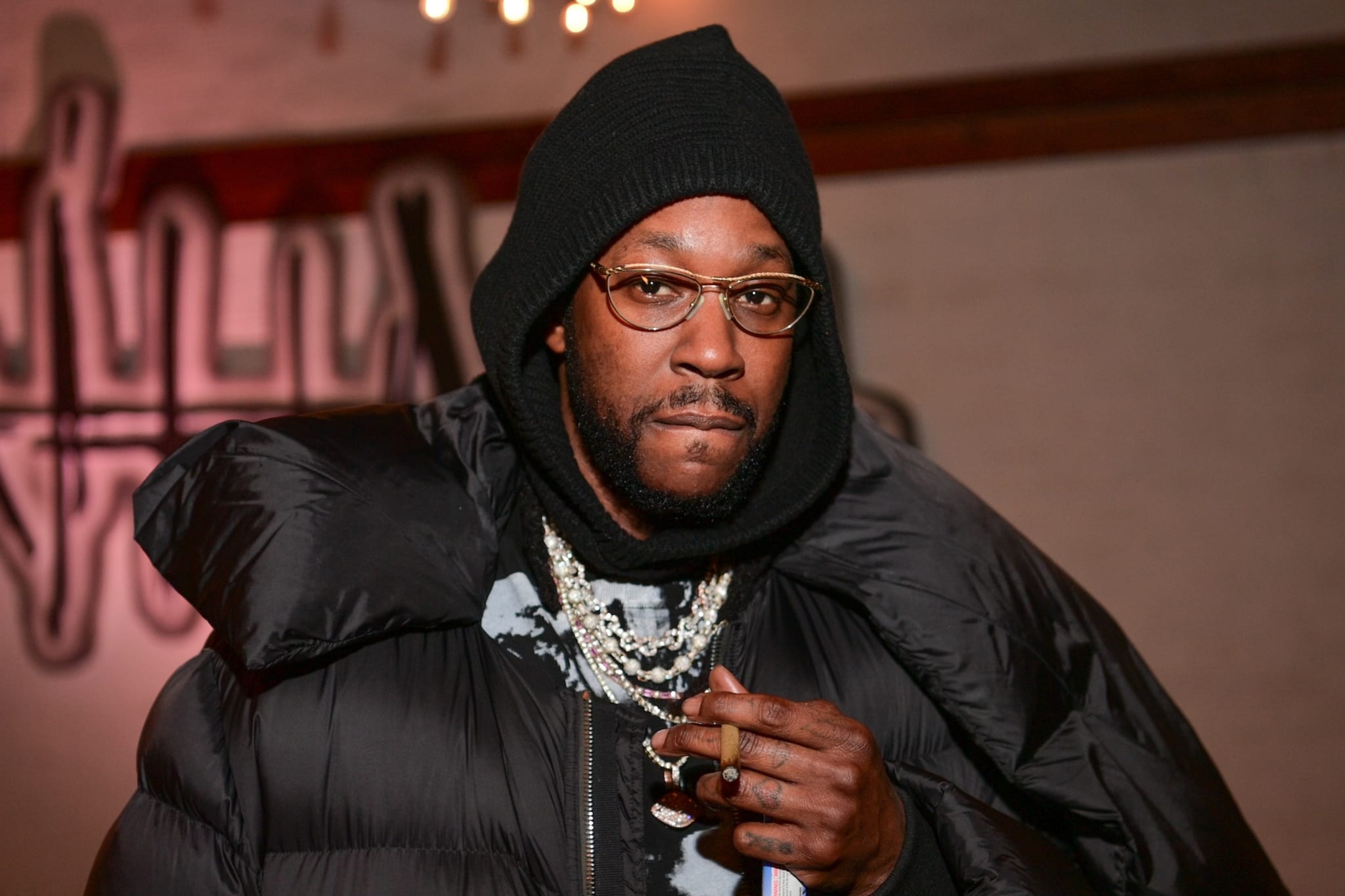 Rapper 2 Chainz wearing glasses, a black hoodie, and a puffy jacket, with layered necklaces, holding a cigar.