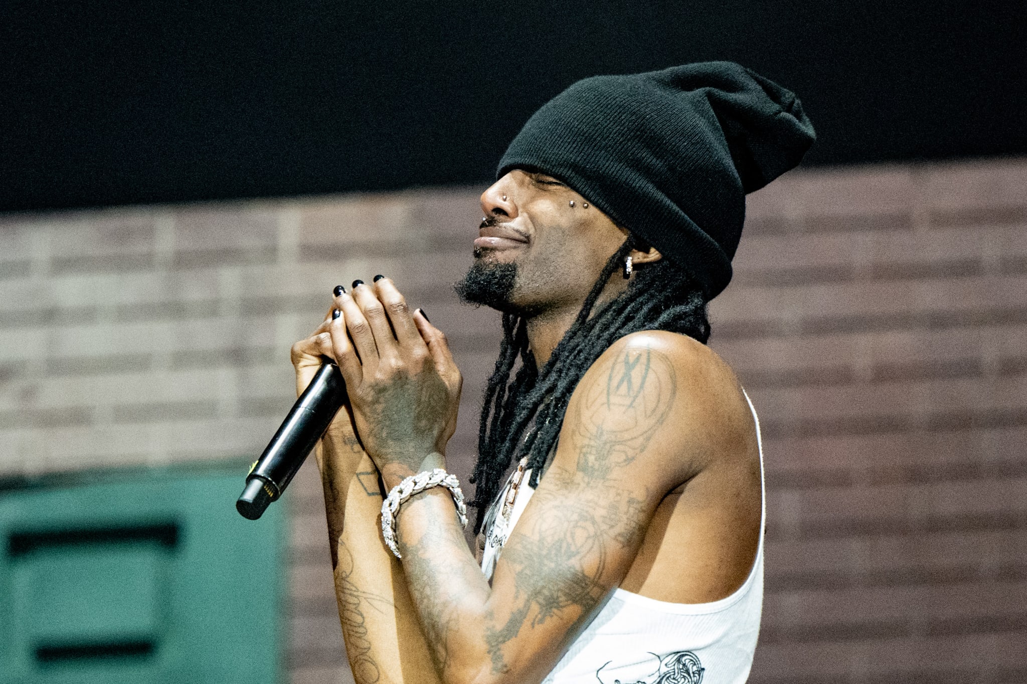 Playboi Carti with braided hair and tattoos, wearing a black beanie and white tank top, holds a microphone on stage.