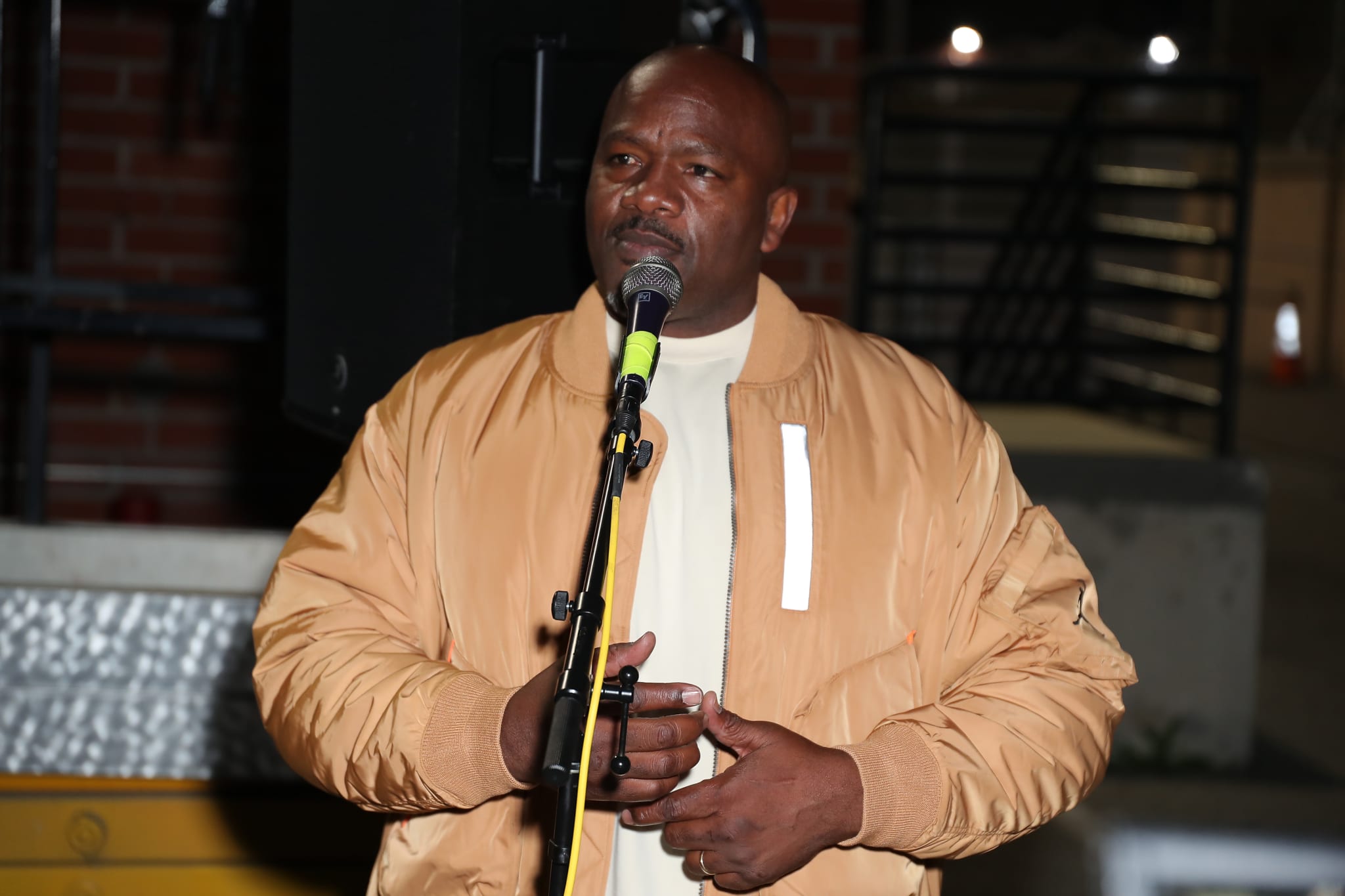A man in a tan jacket speaks into a microphone at an outdoor event. The background is dimly lit.