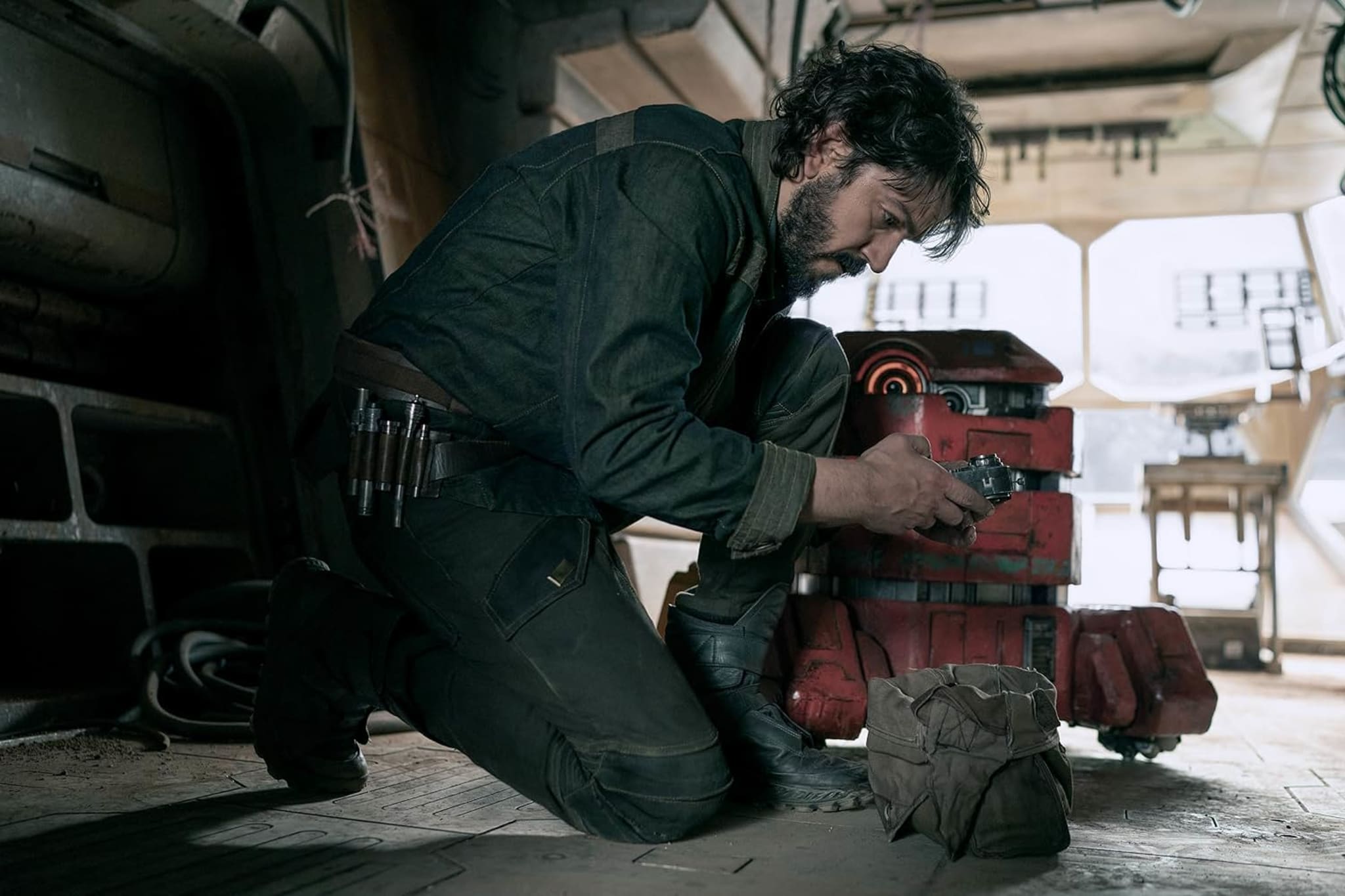 A man kneels in a dimly lit room, working on a red droid. The setting appears to be a sci-fi environment with mechanical elements.