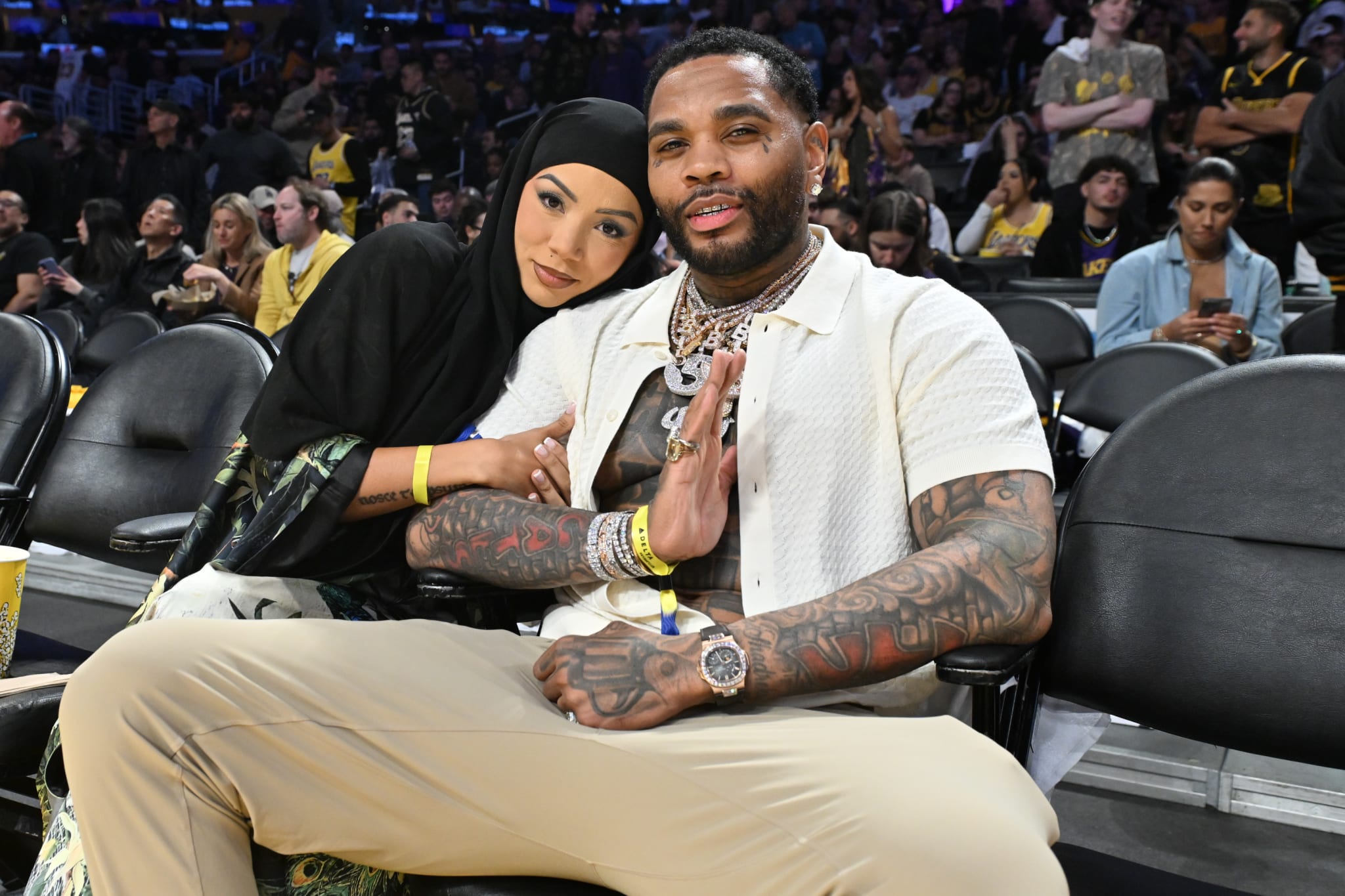 A man with tattoos and a woman in a black hijab sit courtside at a basketball game, posing for a photo.
