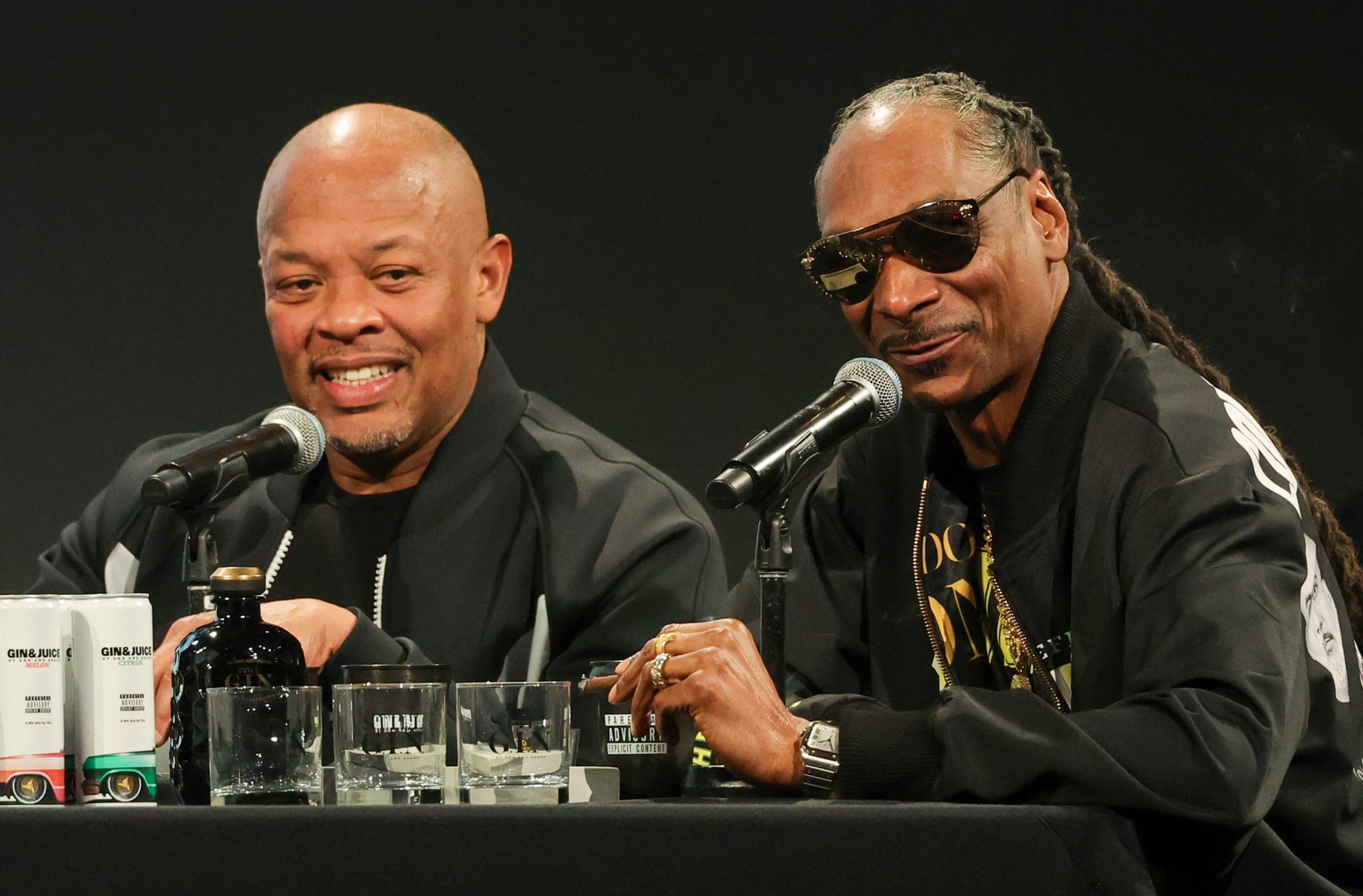 Dr. Dre and Snoop Dogg speaking at a press event, both wearing black jackets. Dr. Dre is smiling, and Snoop Dogg is wearing sunglasses.