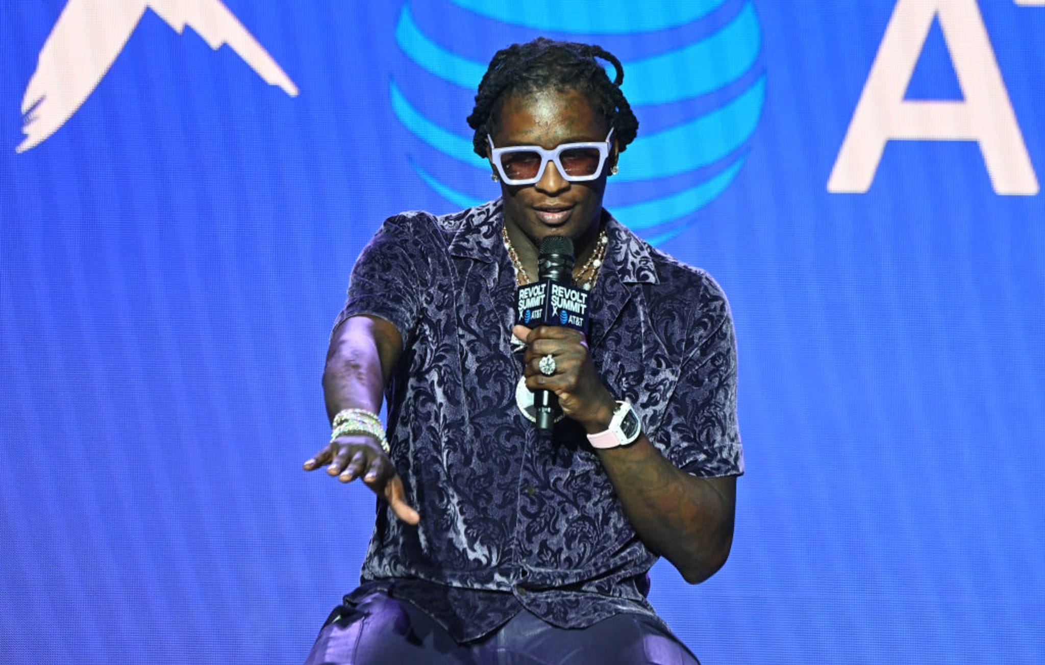 A man wearing sunglasses and a patterned shirt speaks into a microphone on stage, with a blue background featuring logos.