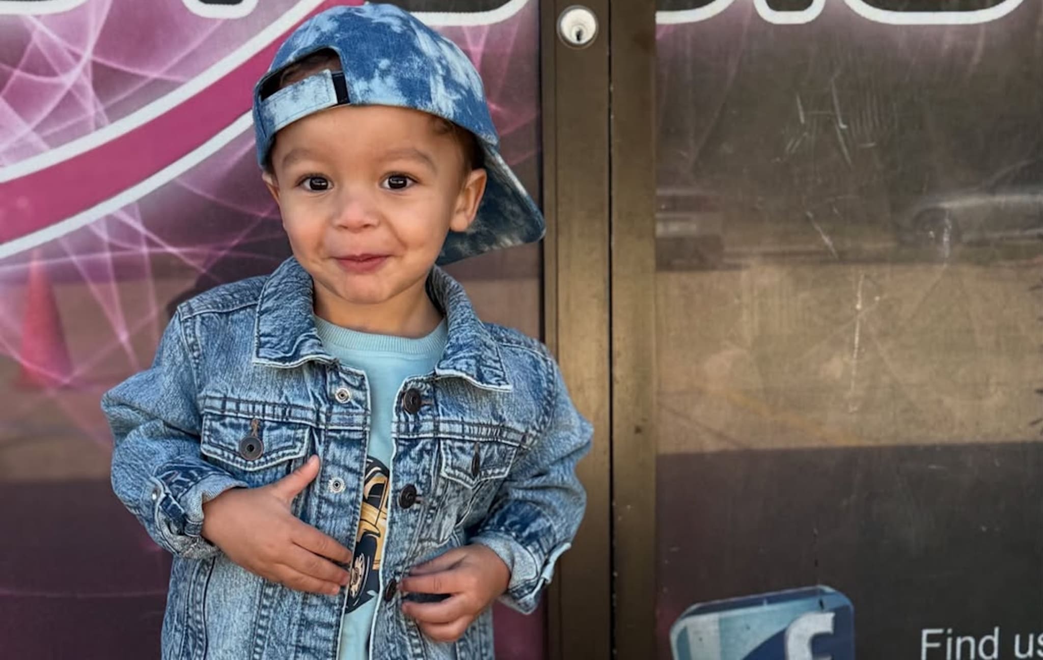 A young child wearing a denim jacket and a blue tie-dye cap, standing in front of a glass door with a playful expression.