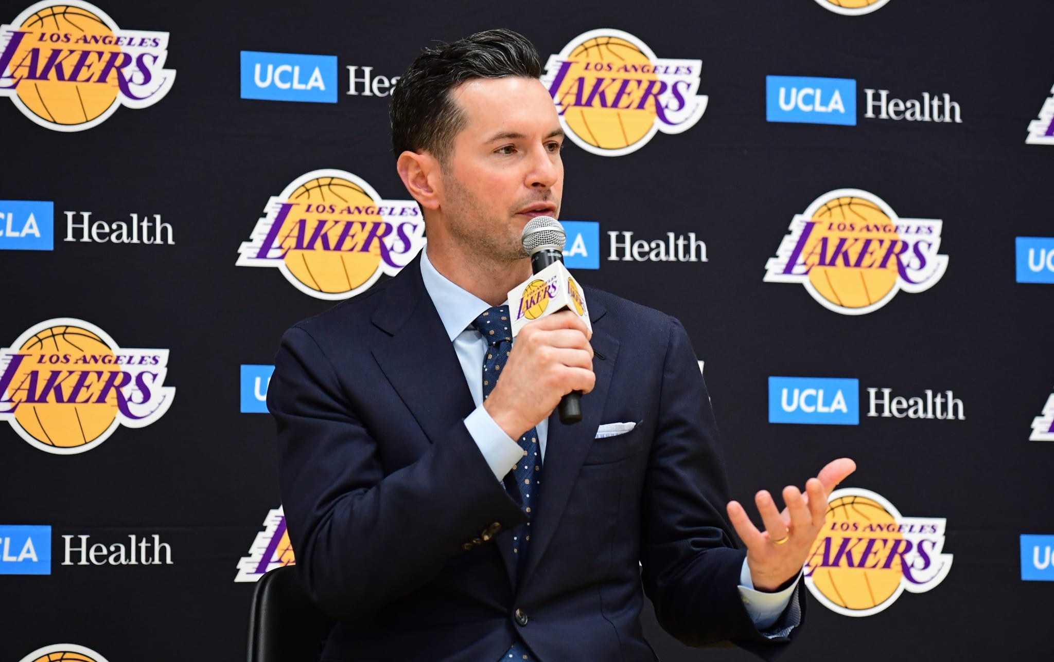 J.J. Redick in a press conference, speaking into a microphone while in a dark suit and tie, seated in front of a Los Angeles Lakers backdrop