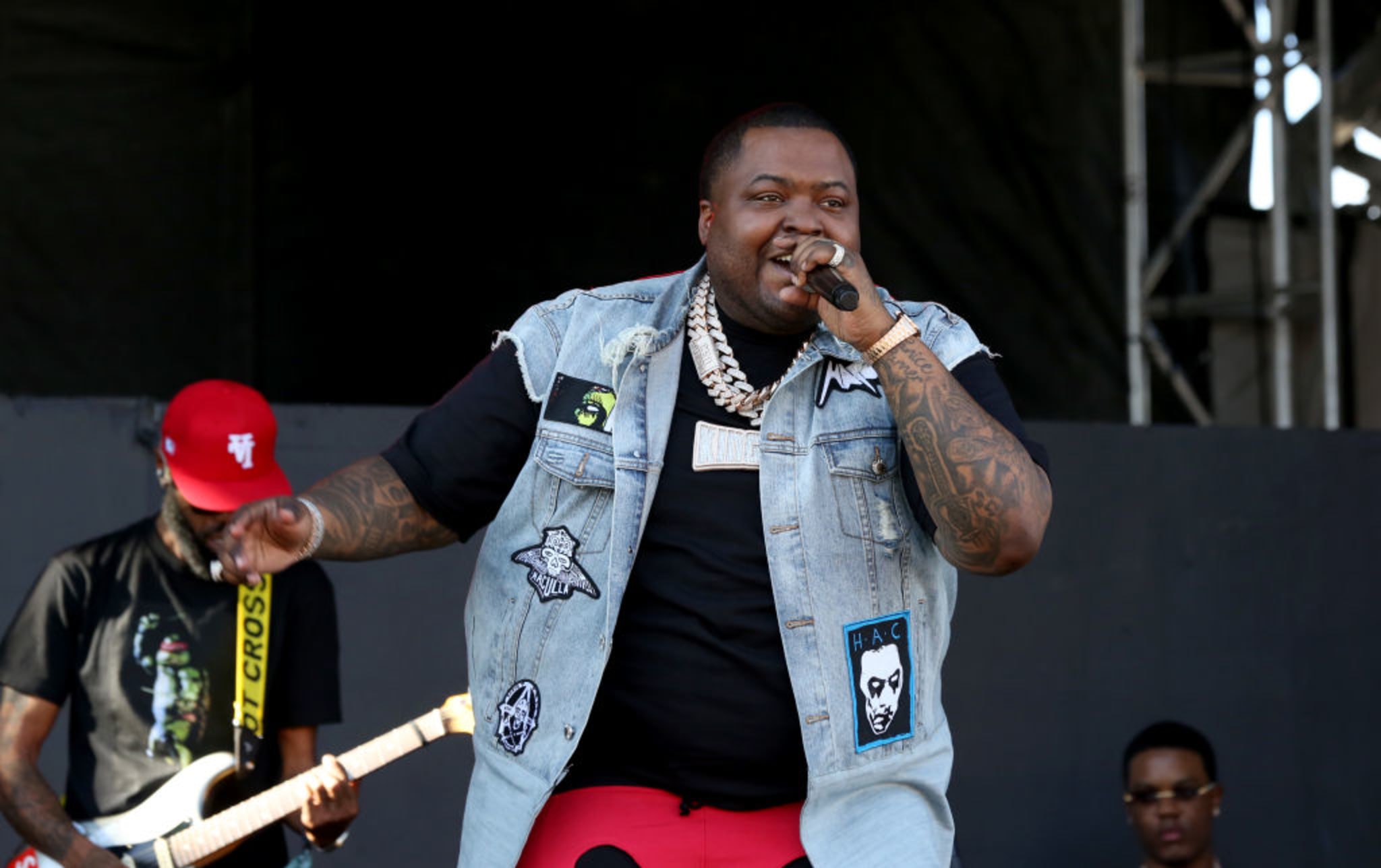 A performer on stage wearing a denim vest and red pants, holding a microphone. A guitarist in a red cap is in the background.