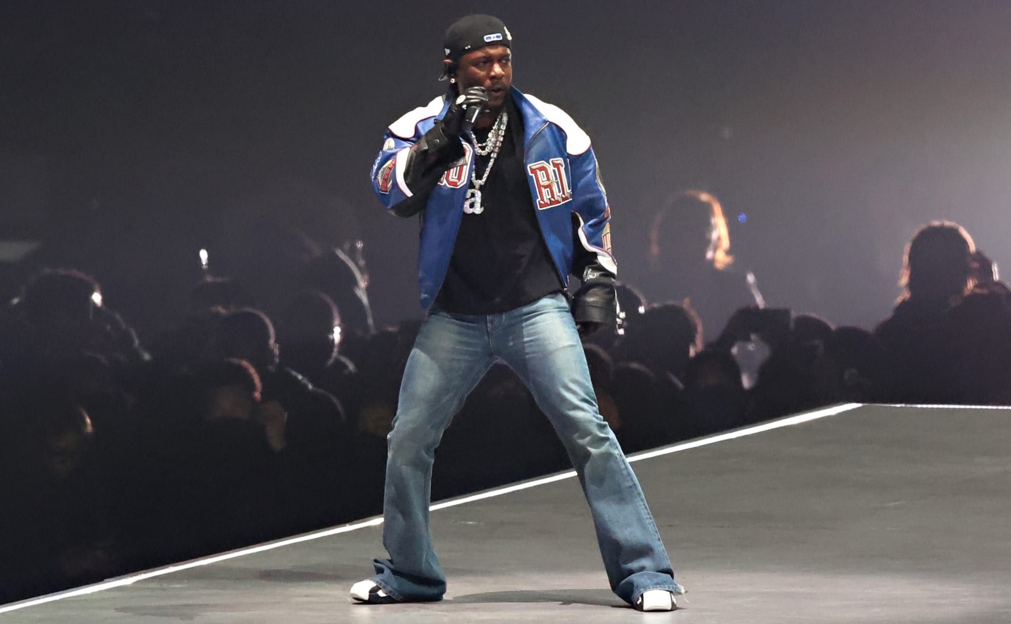 Kendrick Lamar on stage wearing a blue jacket, jeans, and a cap, holding a microphone. The background is dark with an audience visible.