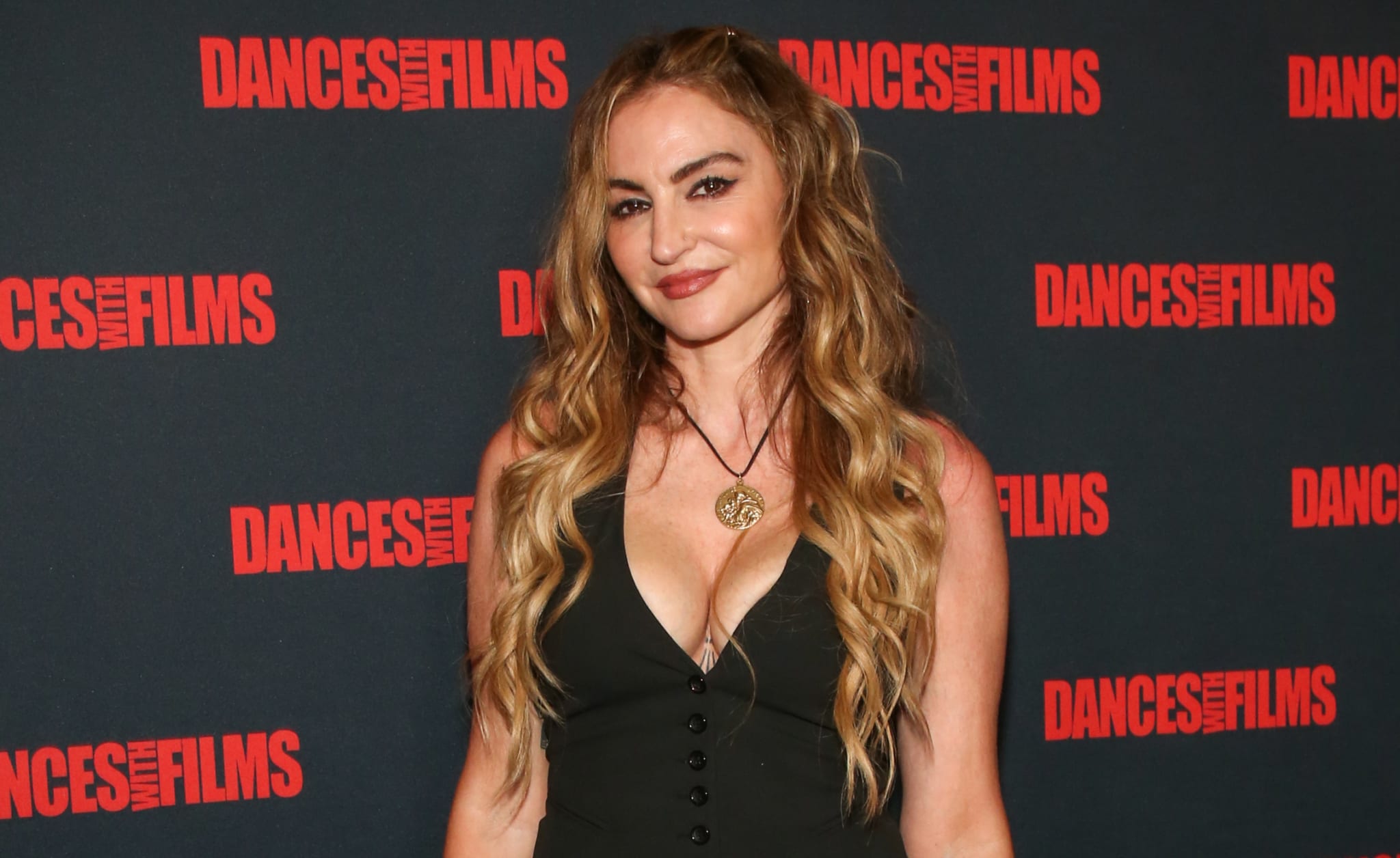 Person smiling at event, wearing a black sleeveless dress with a plunging neckline and a pendant necklace