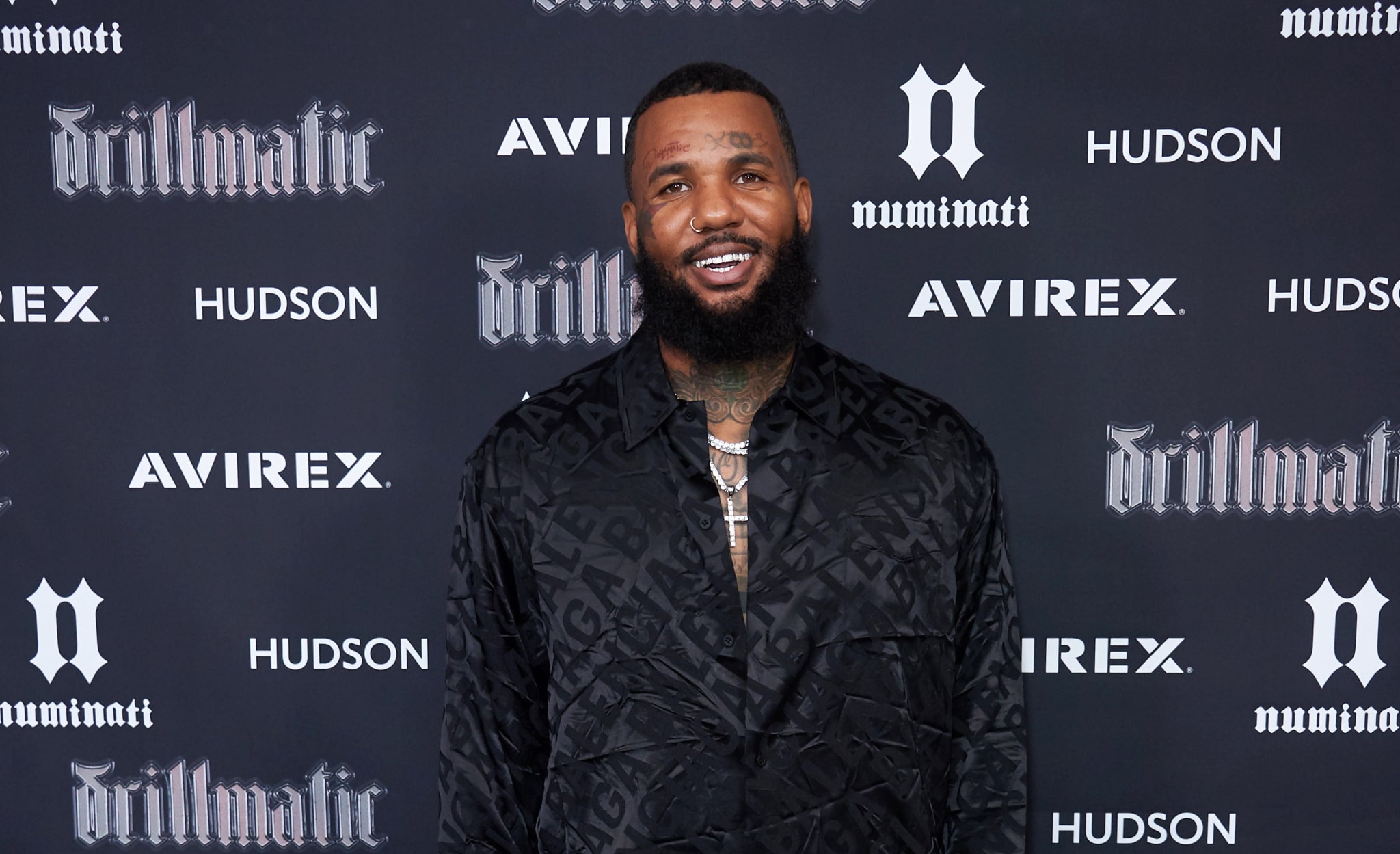 The Game poses on the red carpet in a black patterned suit at a music event, smiling. Logos of Avirex, Hudson, and "drillmatic" are visible in the background
