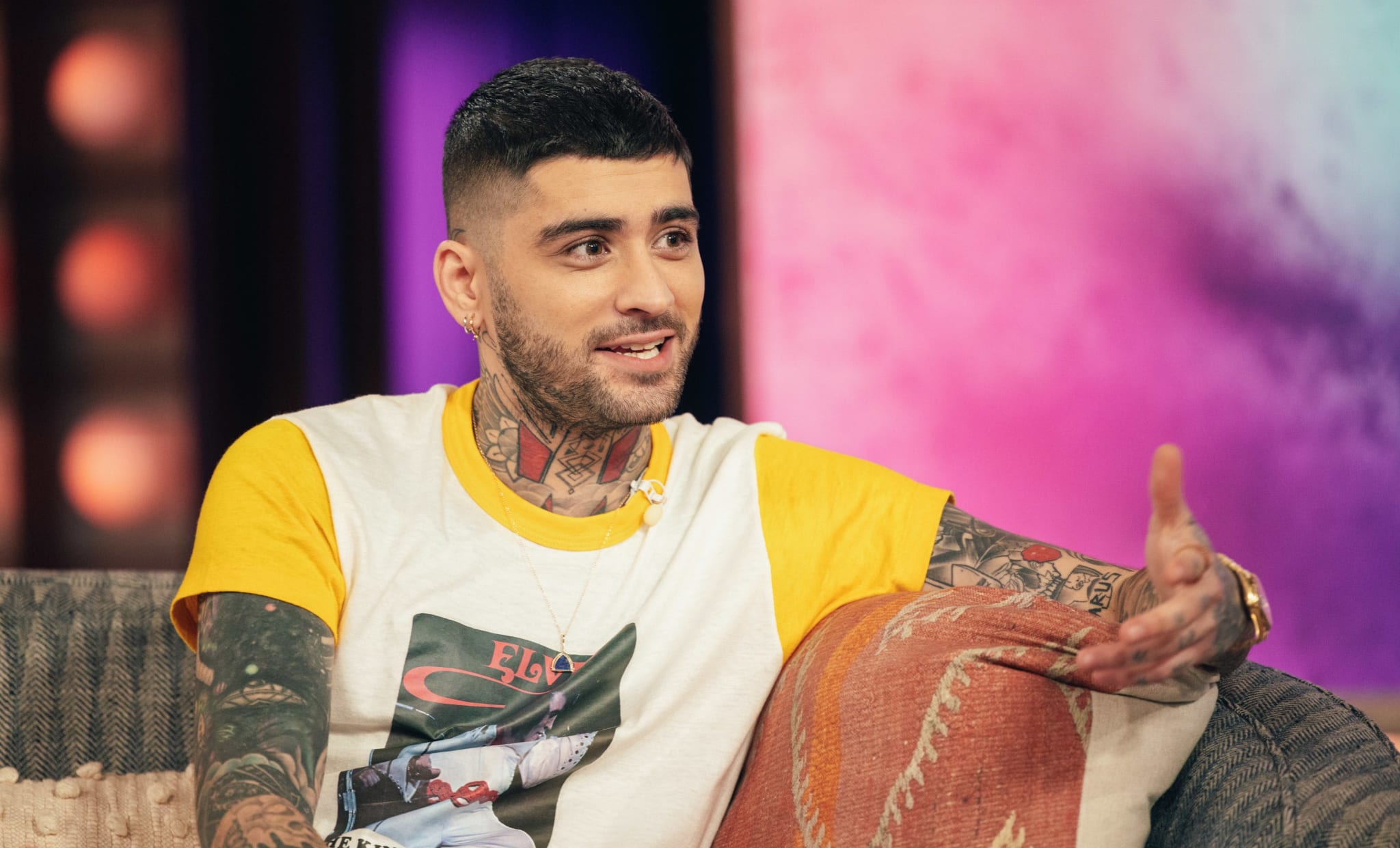 Zayn Malik sitting on a couch, animated while in conversation, wearing a graphic t-shirt and tattoos visible