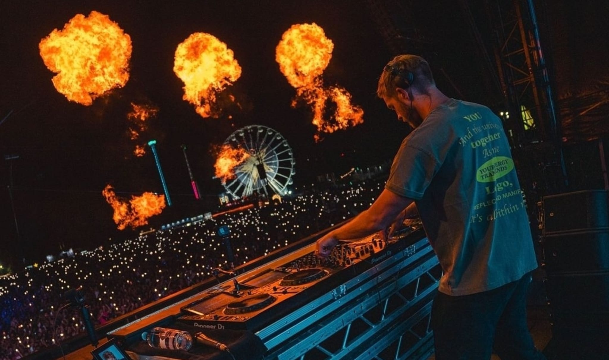 A DJ performs on stage at a music festival, with fire effects burst in the background and a Ferris wheel visible among the crowd of people