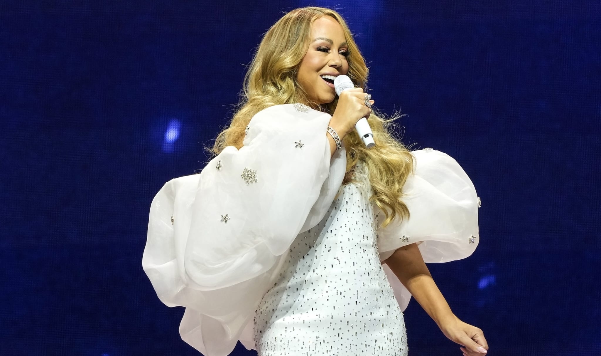 Mariah Carey performing on stage, wearing a white dress with large sleeves, holding a microphone and smiling.