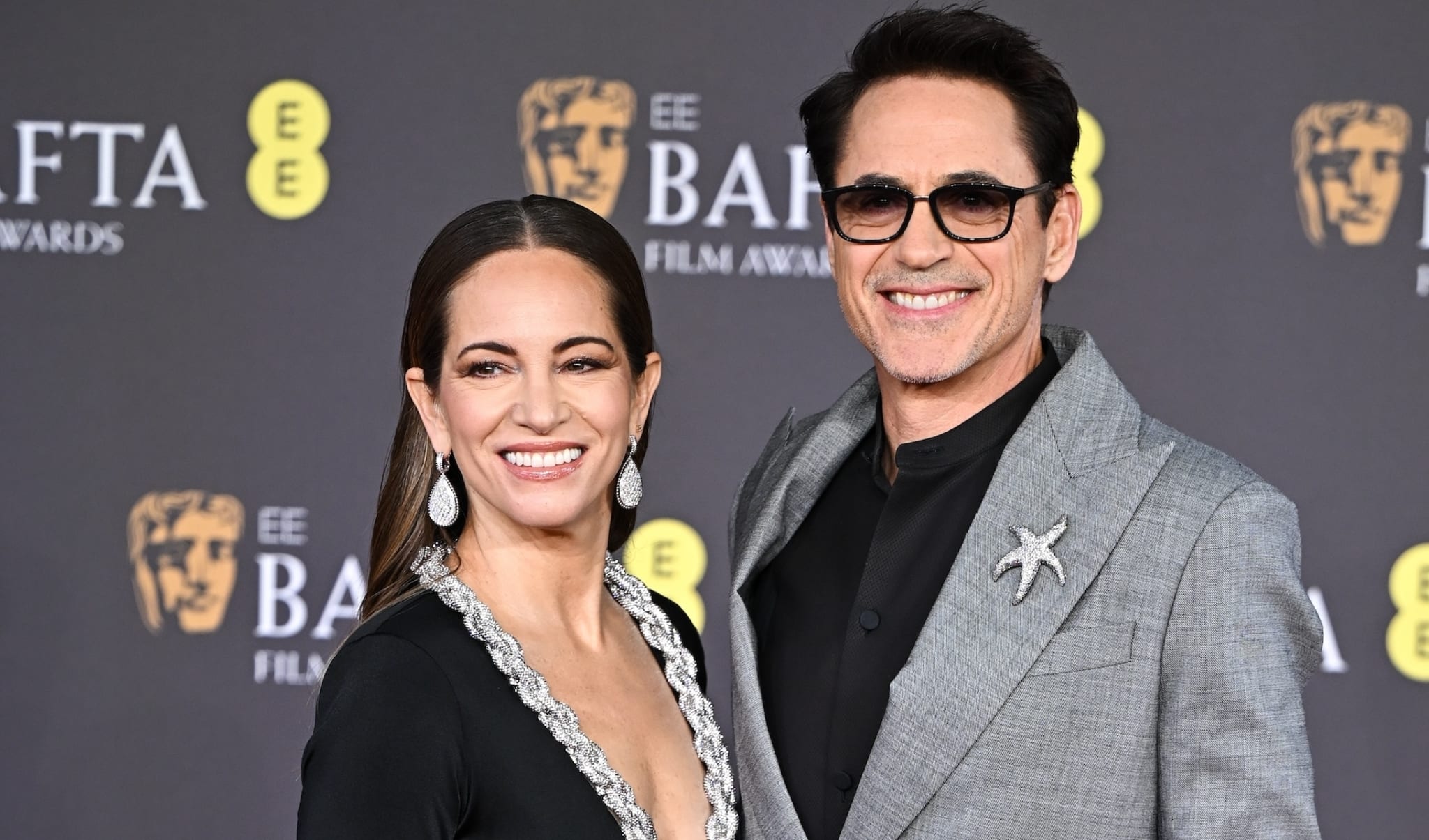 Two people posing at the BAFTA awards, one in a dark V-neck dress and the other in a light suit with glasses