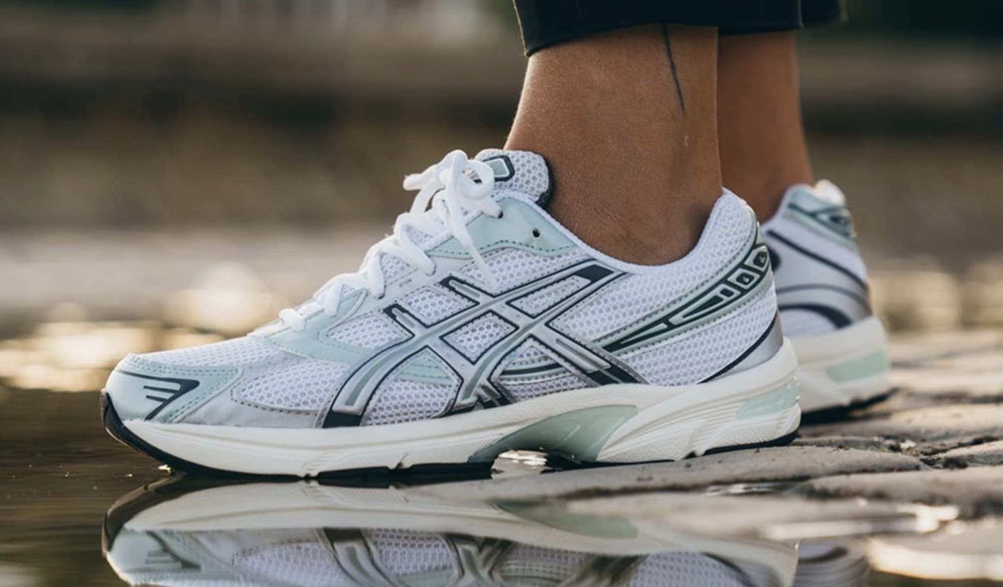 Close-up of a person's legs in black pants wearing Asics sneakers by a reflective puddle on a cobblestone surface