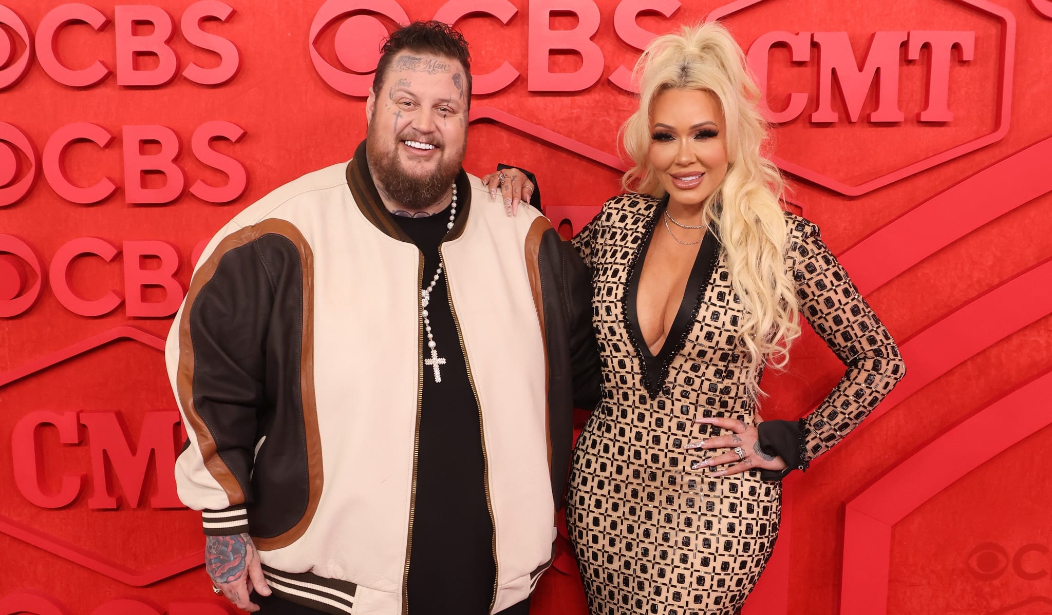 Two people posing together on the red carpet; one is wearing a bomber jacket and the other is in a sequined outfit