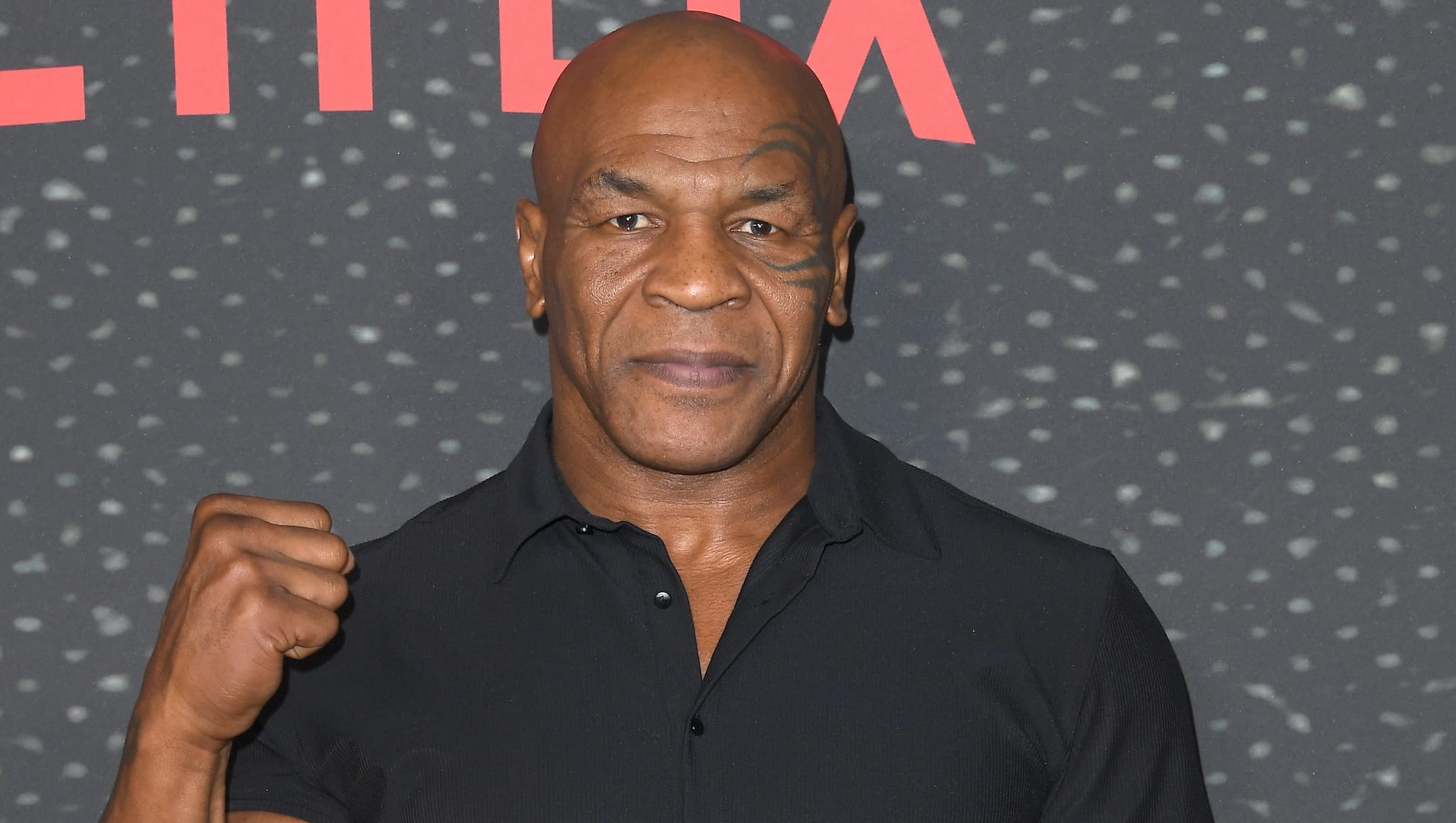 Mike Tyson, wearing a black shirt, stands with a fist raised in front of a speckled background.