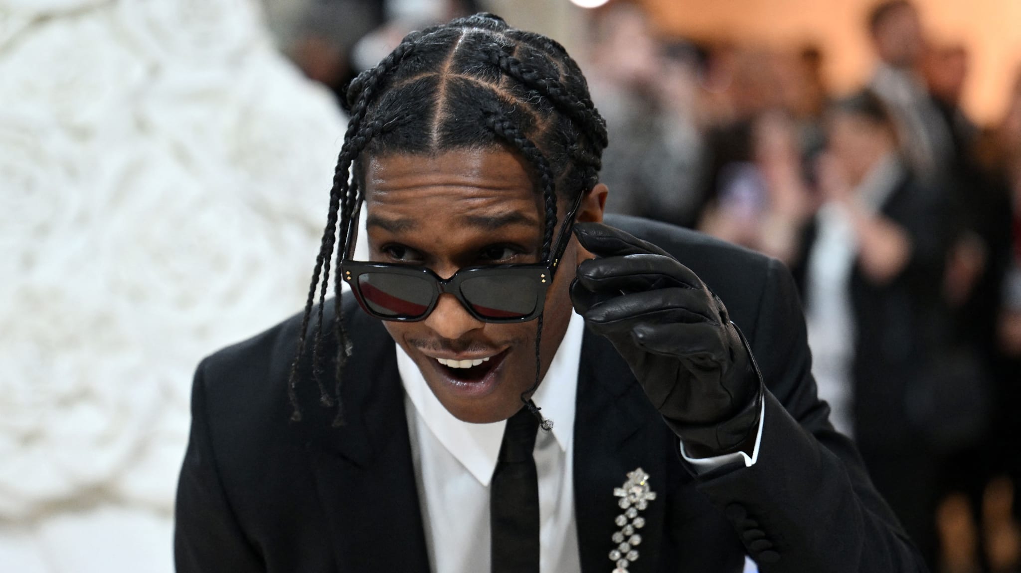 ASAP Rocky, wearing a stylish black suit with a sparkling brooch, black gloves, and dark sunglasses, looks into the camera at a music event
