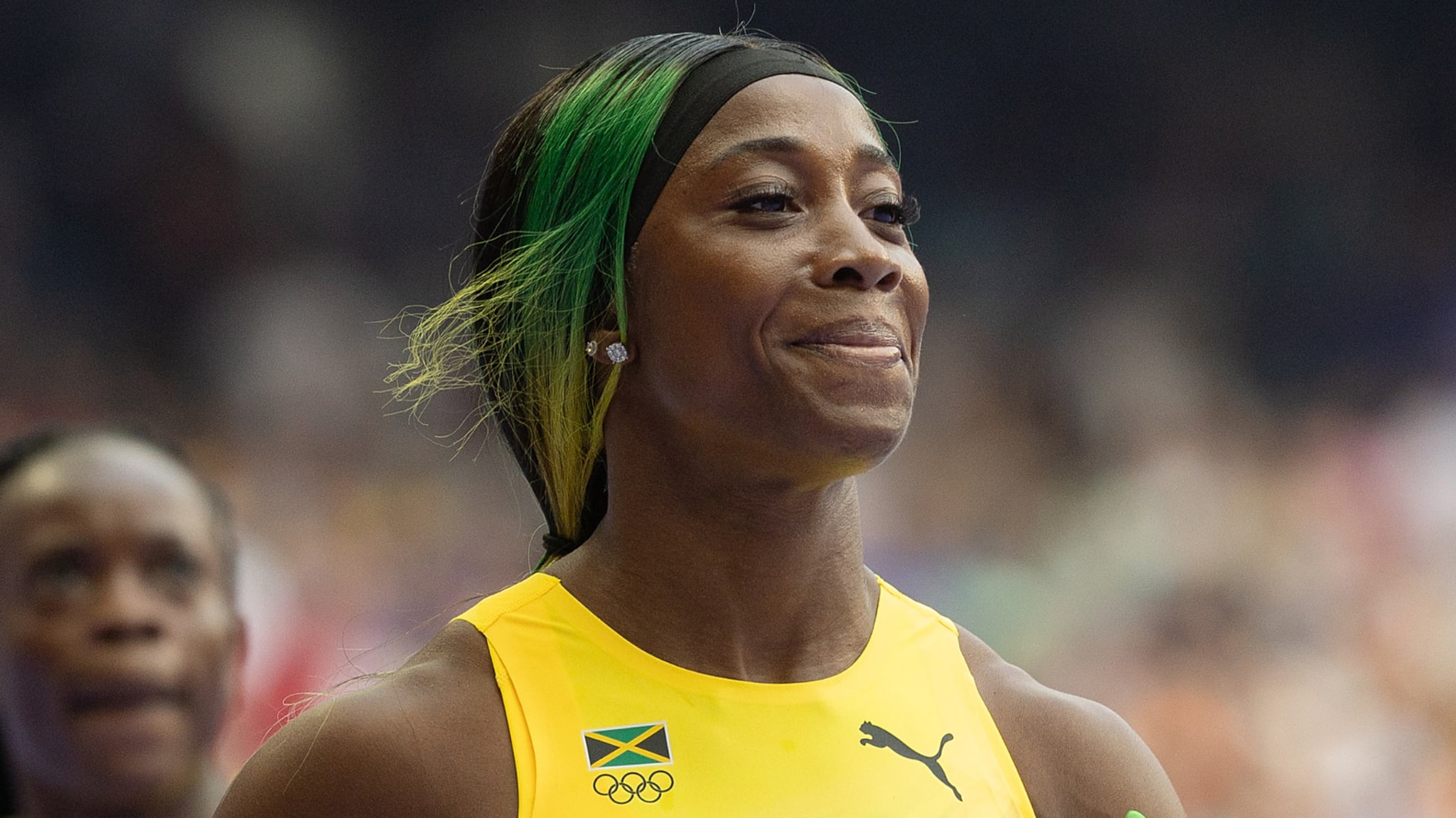 Shelly-Ann Fraser-Pryce is smiling while wearing an athletic outfit at an event. Another person is visible in the background