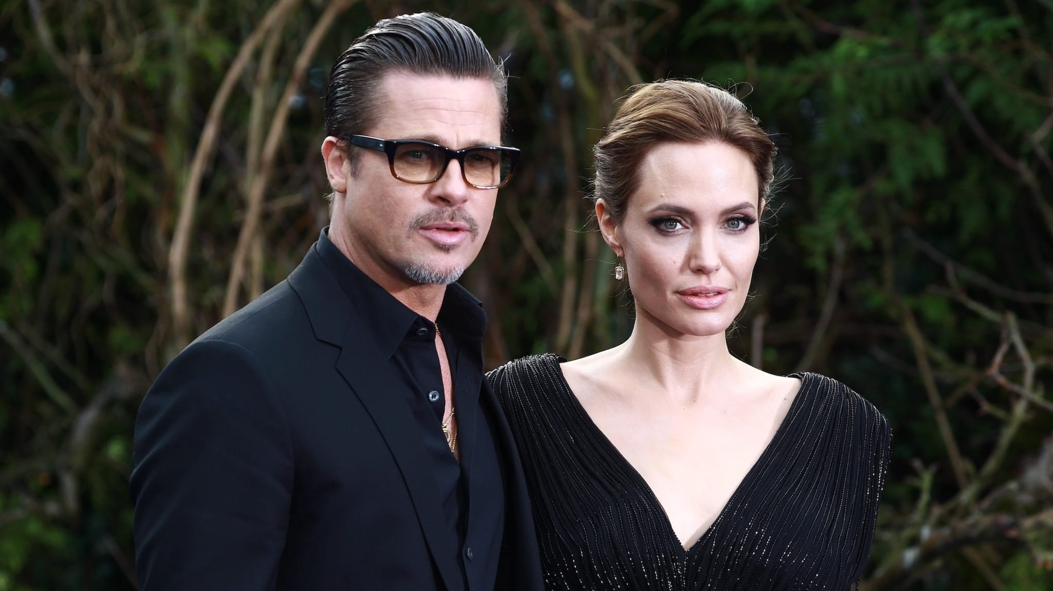 Brad Pitt, wearing a black suit and glasses, stands beside Angelina Jolie, who is dressed in a black, v-neck gown