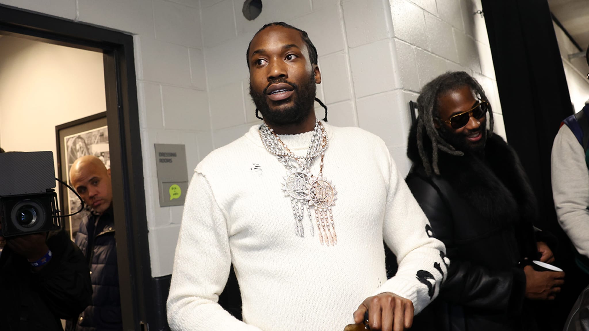 Meek Mill wearing a white sweater with intricate chain necklaces, walks backstage, accompanied by a man in a black coat and sunglasses