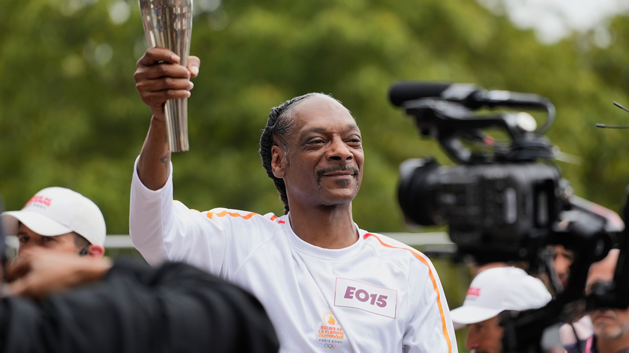 Snoop Dogg holds a torch while wearing a white sports outfit surrounded by people and a camera crew