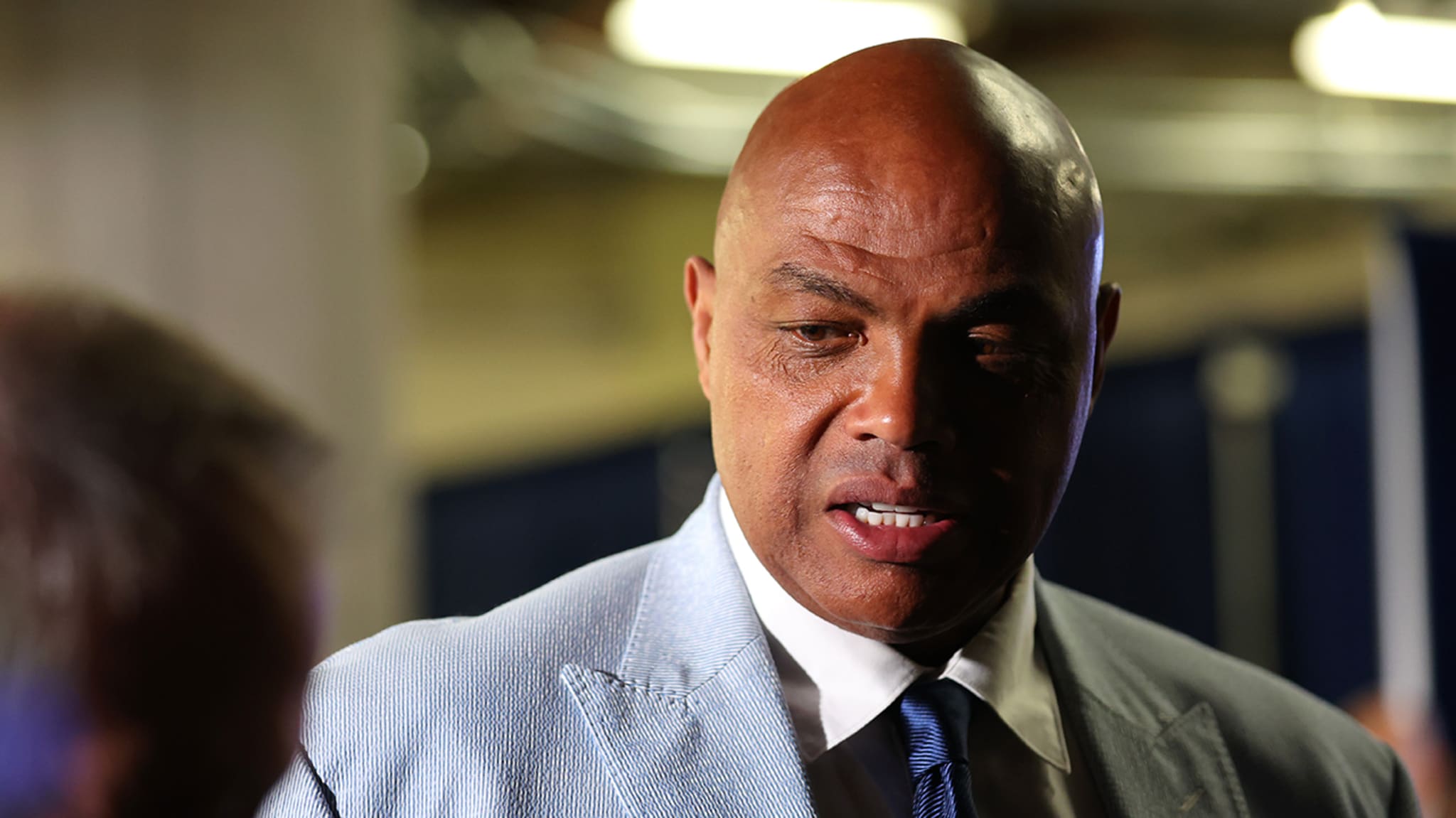 Charles Barkley in a light suit and tie, speaking with someone at a sports event