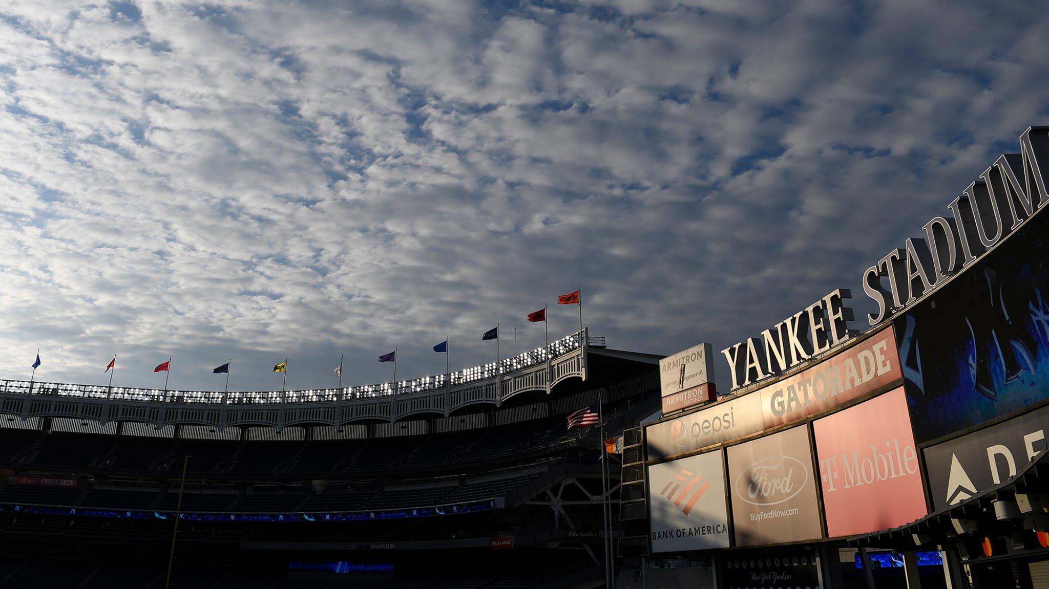 Yankee Stadium sign with a partly cloudy sky above, flags lined up atop the stadium. Stadium lights and sponsor banners visible