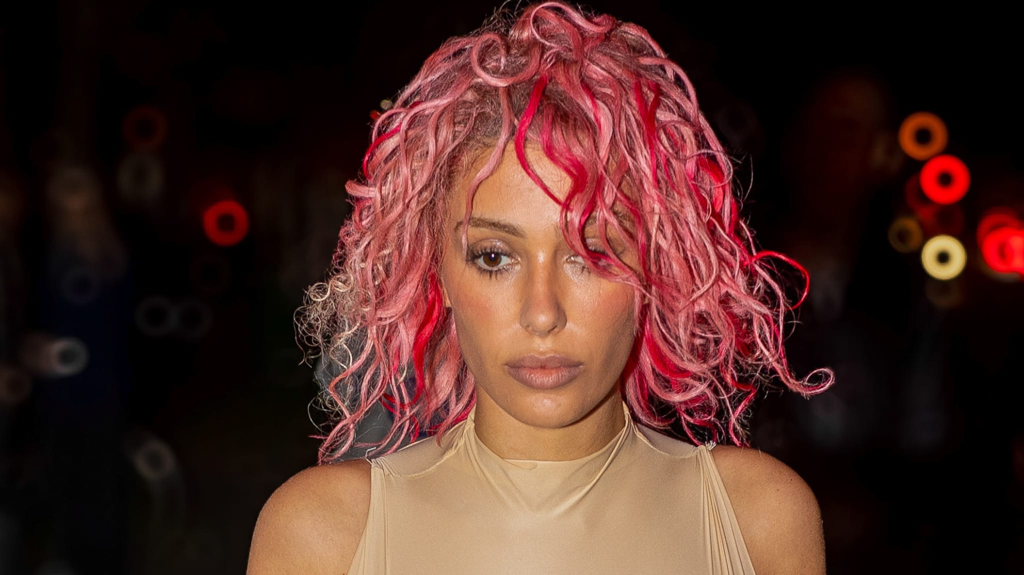A woman with curly hair styled in tight curls. She is wearing a sleeveless, fitted outfit with a high neckline. The background is dark with blurred lights