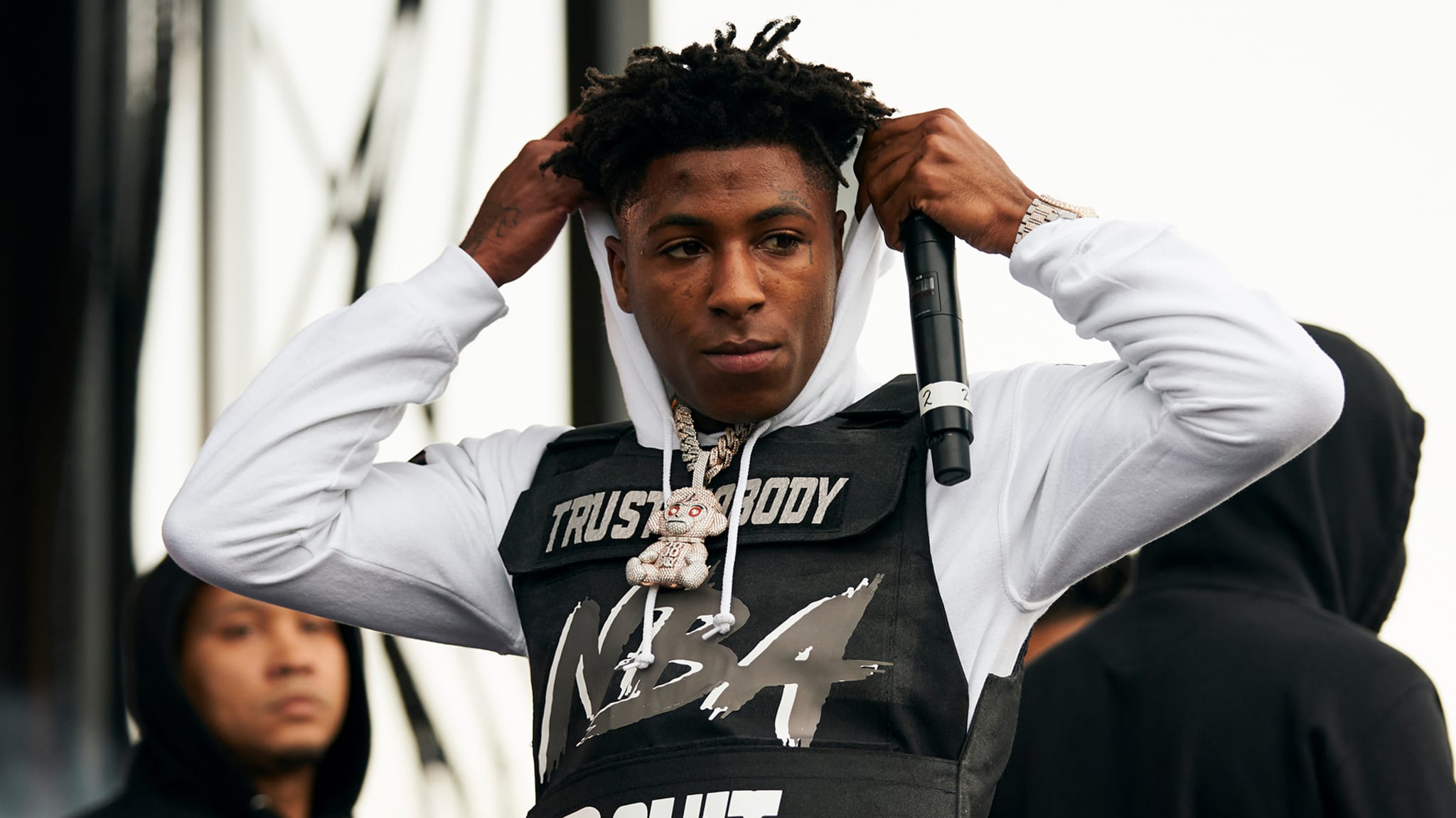 Rapper NBA YoungBoy on stage, wearing a white hoodie under a black vest with "Trust Nobody" and "NBA" printed on it, holding a microphone