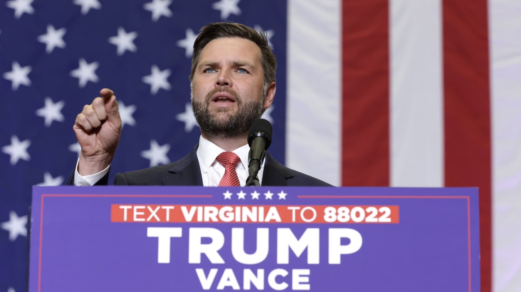 J.D. Vance speaks at a podium with "TEXT VIRGINIA TO 88022 TRUMP VANCE" in front of a large American flag backdrop