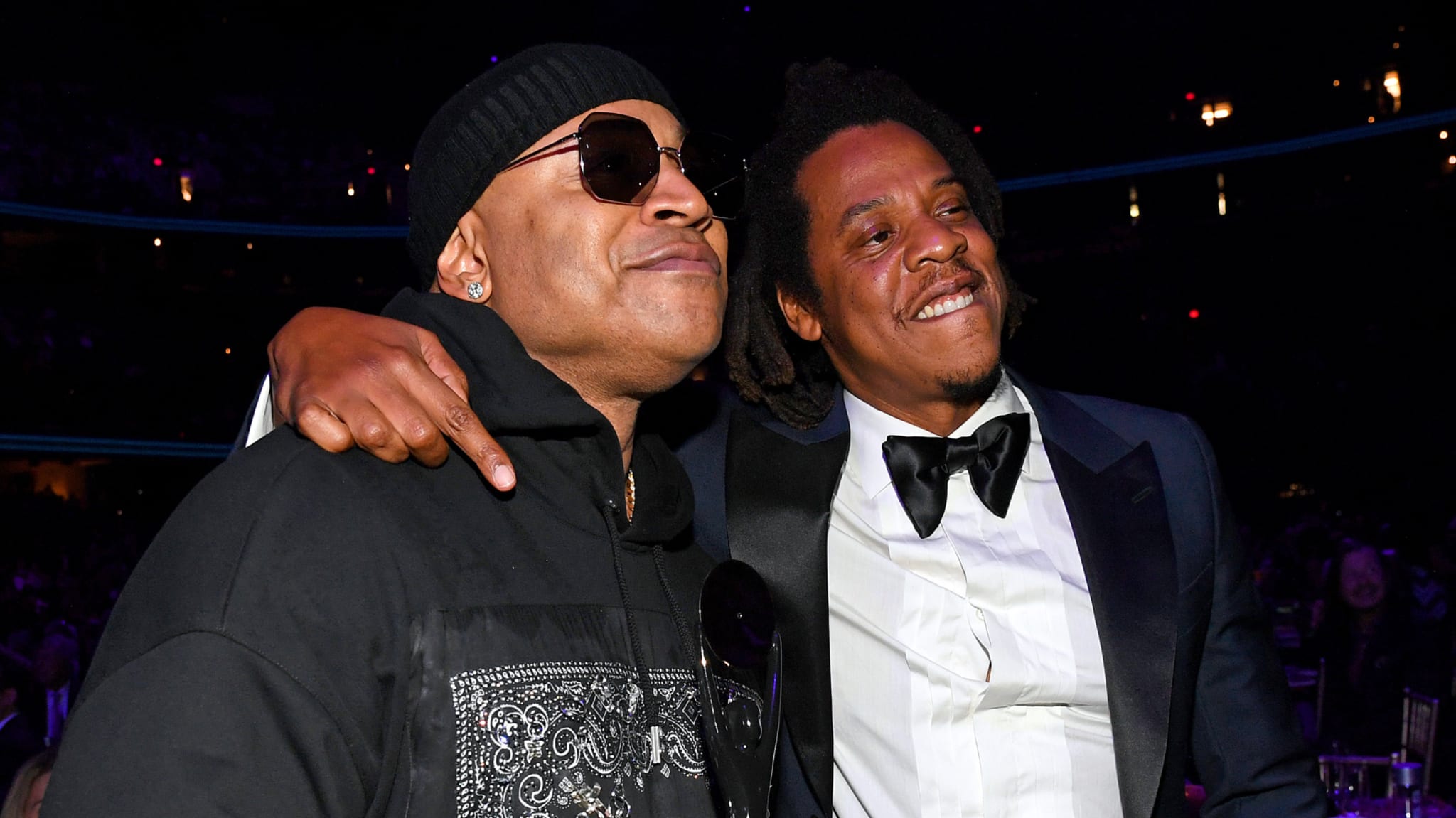 LL Cool J in a black hoodie and beanie, and Jay-Z in a black tuxedo with a bow tie, are smiling and embracing at a music event