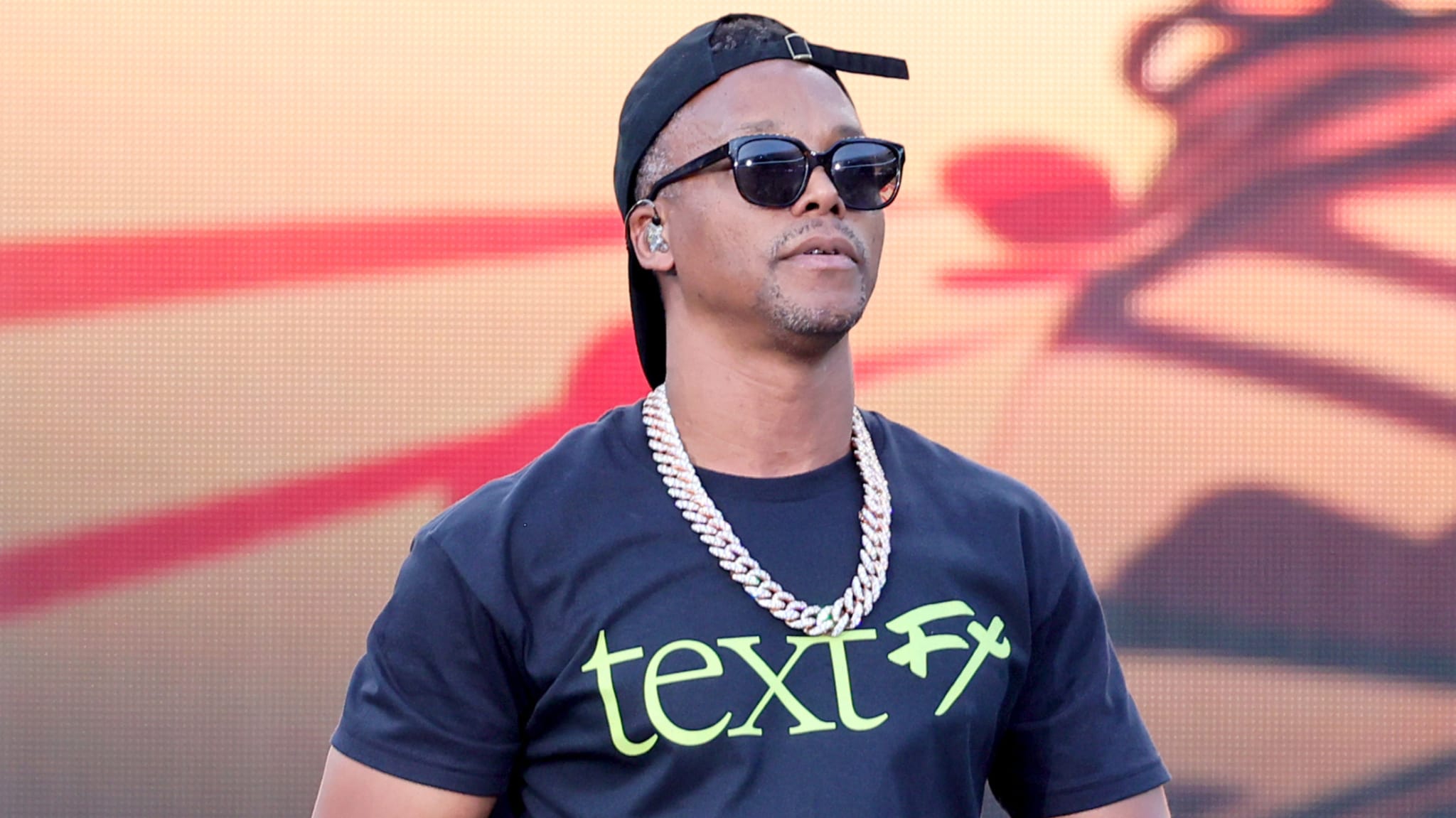 Lupe Fiasco wearing sunglasses, a backwards cap, a chain necklace, and a t-shirt with "text FX" printed on it, at a music event