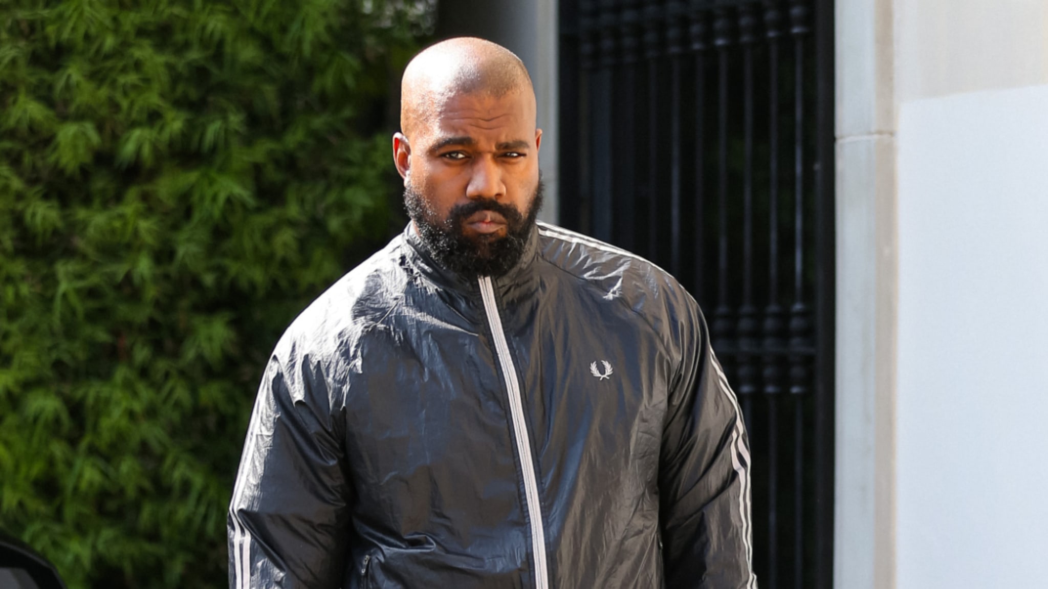 Kanye West wearing a zip-up jacket and looking towards the camera with a serious expression. Bushes and a building are visible in the background