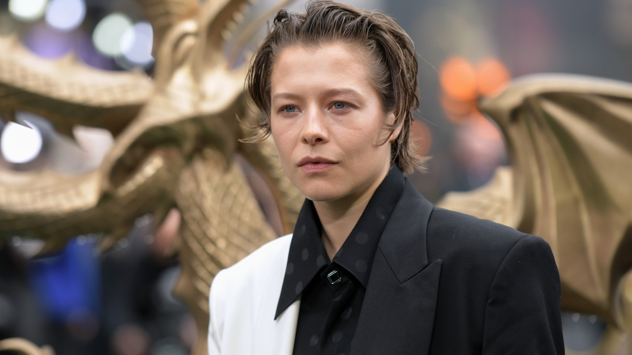 Individual posing on a red carpet at an event, wearing a stylish half black, half white suit jacket with a dragon statue in the background