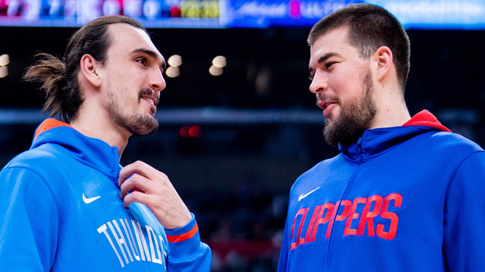 Two basketball players in team attire, one in a "Thunder" hoodie and the other in a "Clippers" hoodie, having a conversation on the court