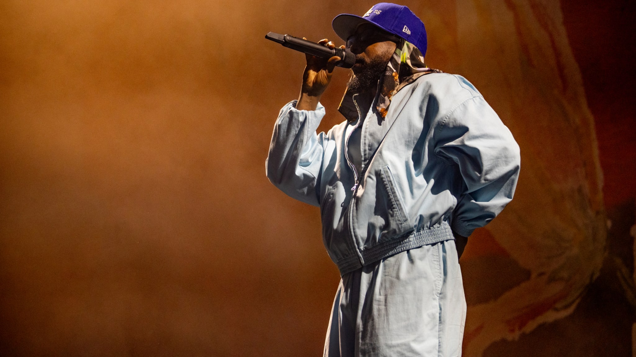Kendrick performing on stage, wearing a loose-fitting jumpsuit and a baseball cap, holding a microphone close to her mouth