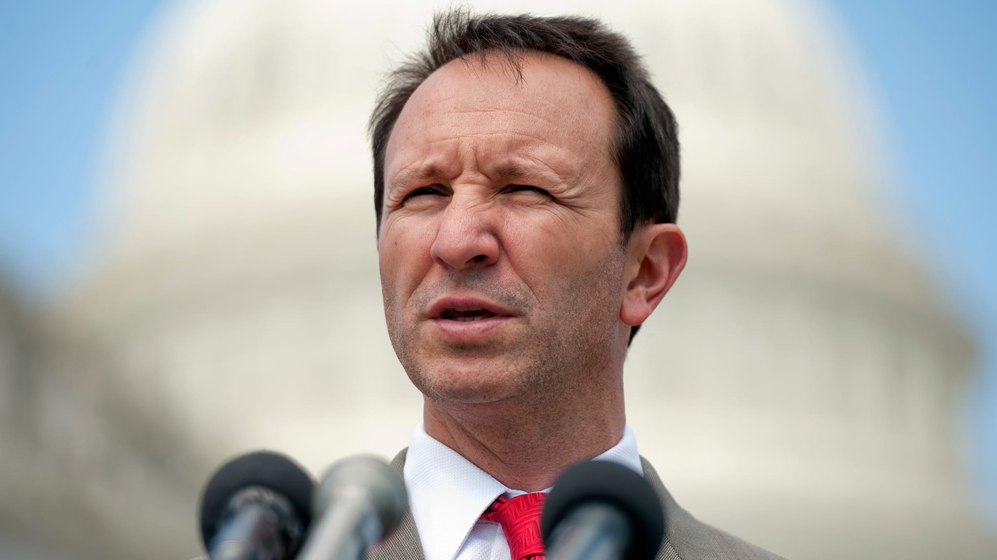 A man in a suit and red tie speaks in front of the U.S. Capitol building, with multiple microphones positioned in front of him