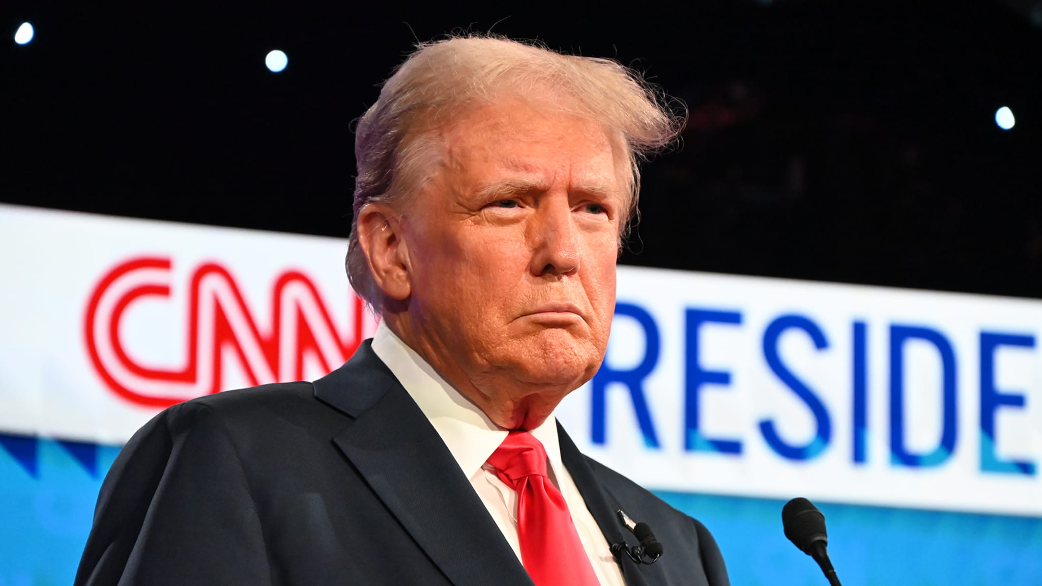 Donald Trump stands in front of a backdrop with the CNN logo and the word "President." He is wearing a suit with a white shirt and red tie