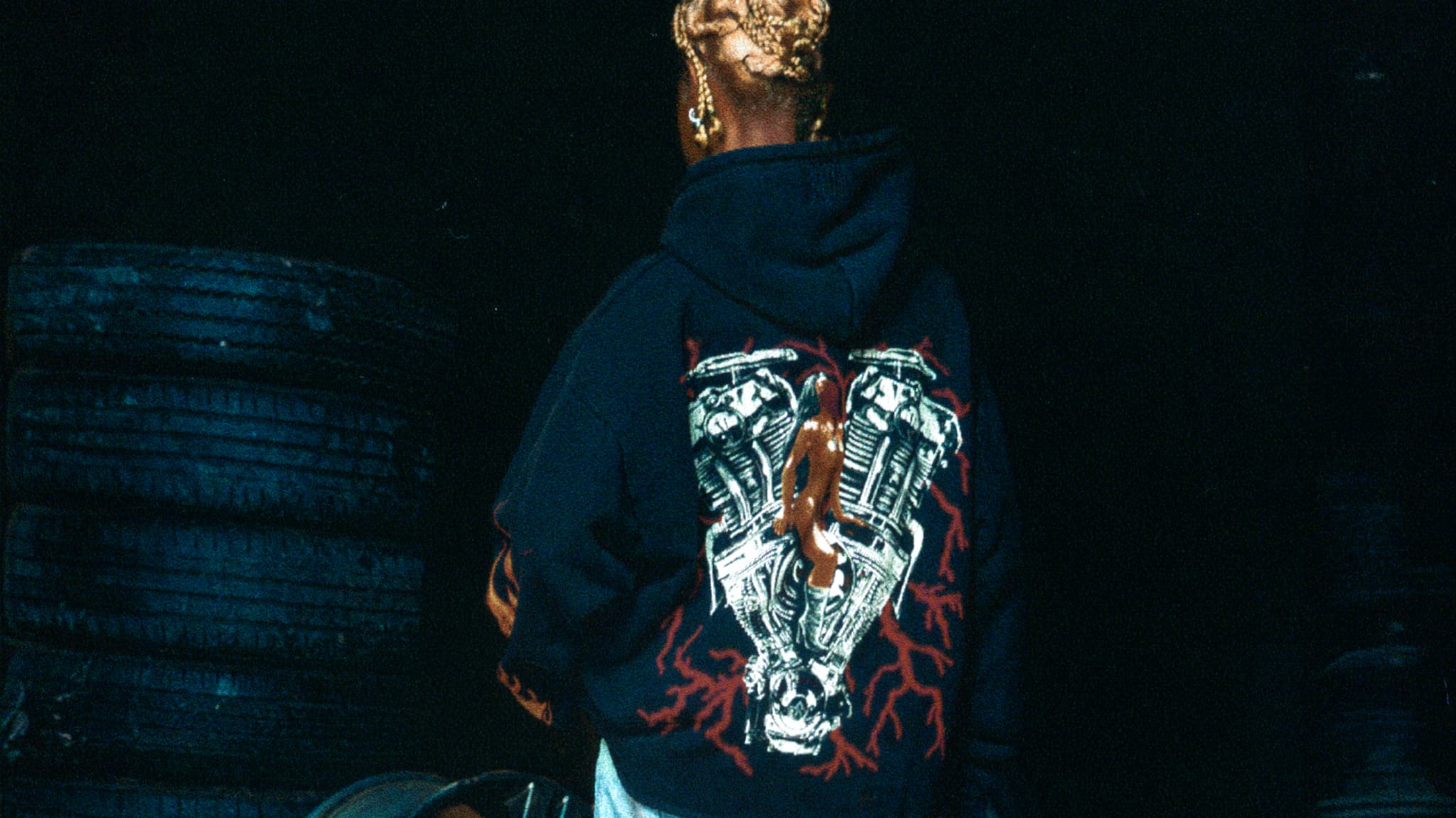 Person with intricate hair braid, wearing a hoodie featuring a large design of an engine and red accents on the back, stands in front of stacked tires