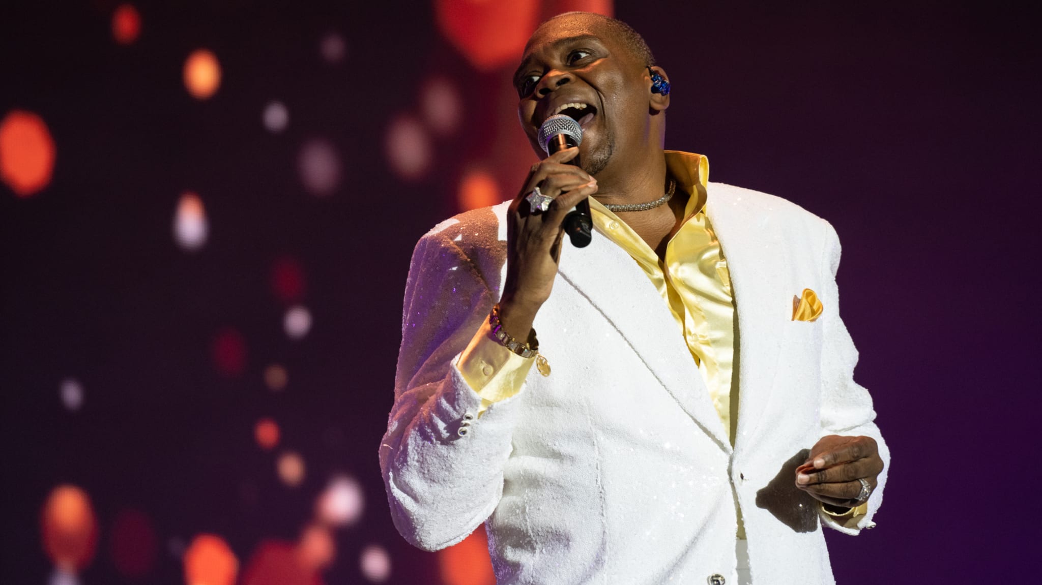 A singer wearing a white suit with a yellow shirt performs onstage, holding a microphone against a backdrop of glowing lights