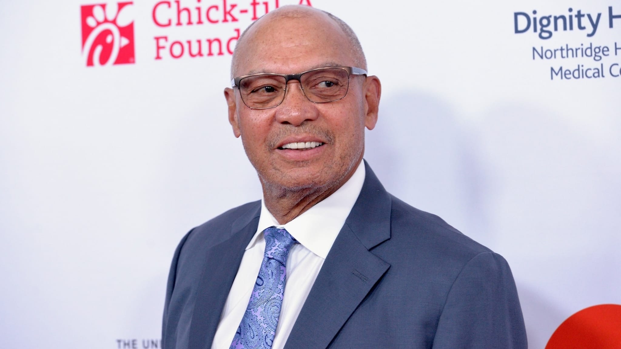 An unidentified person in a suit and tie poses in front of a backdrop with logos for Chick-fil-A Foundation and Dignity Health
