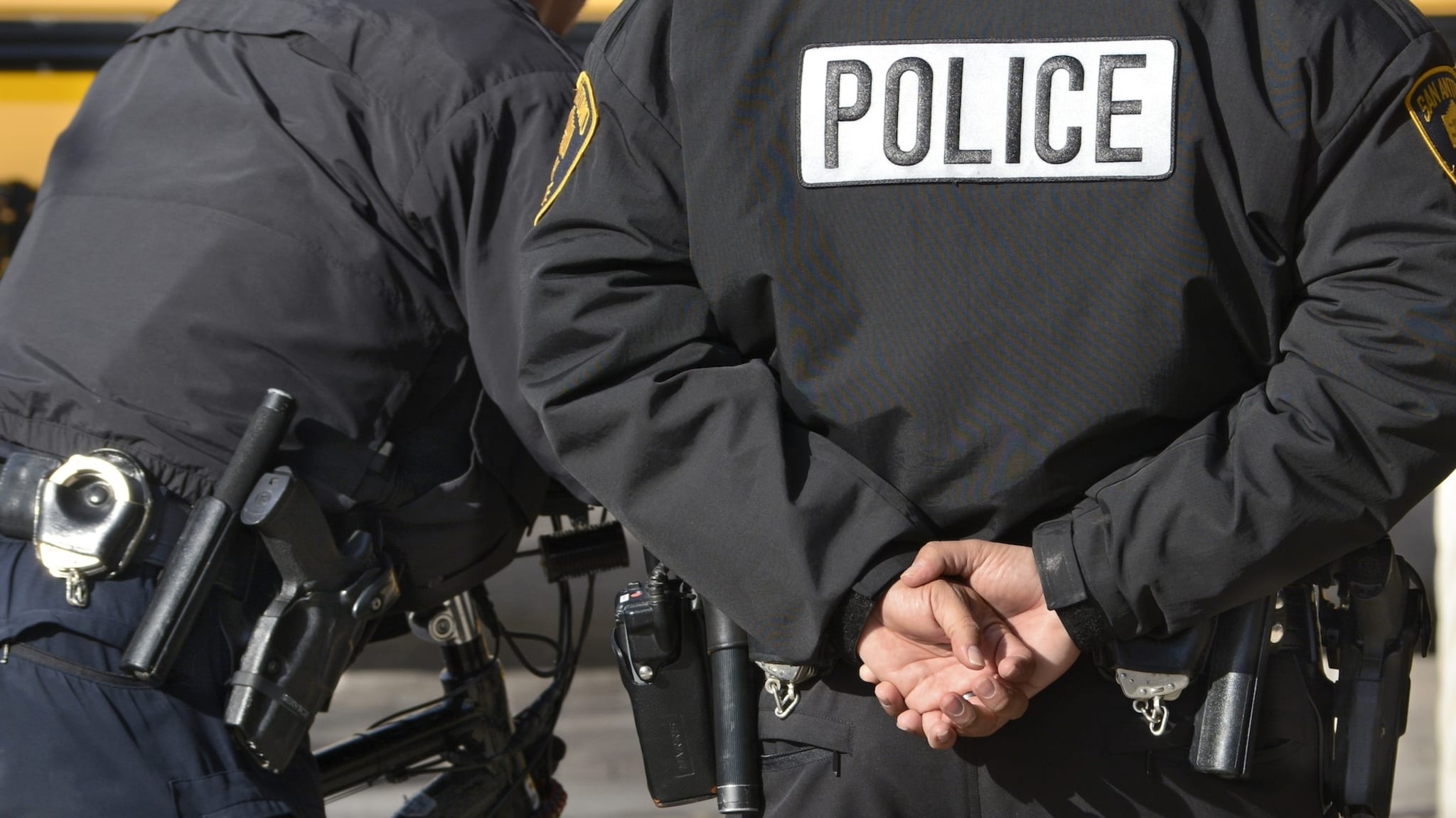 Two police officers, one with their hands behind their back. They are in black police uniforms with visible badges and equipment