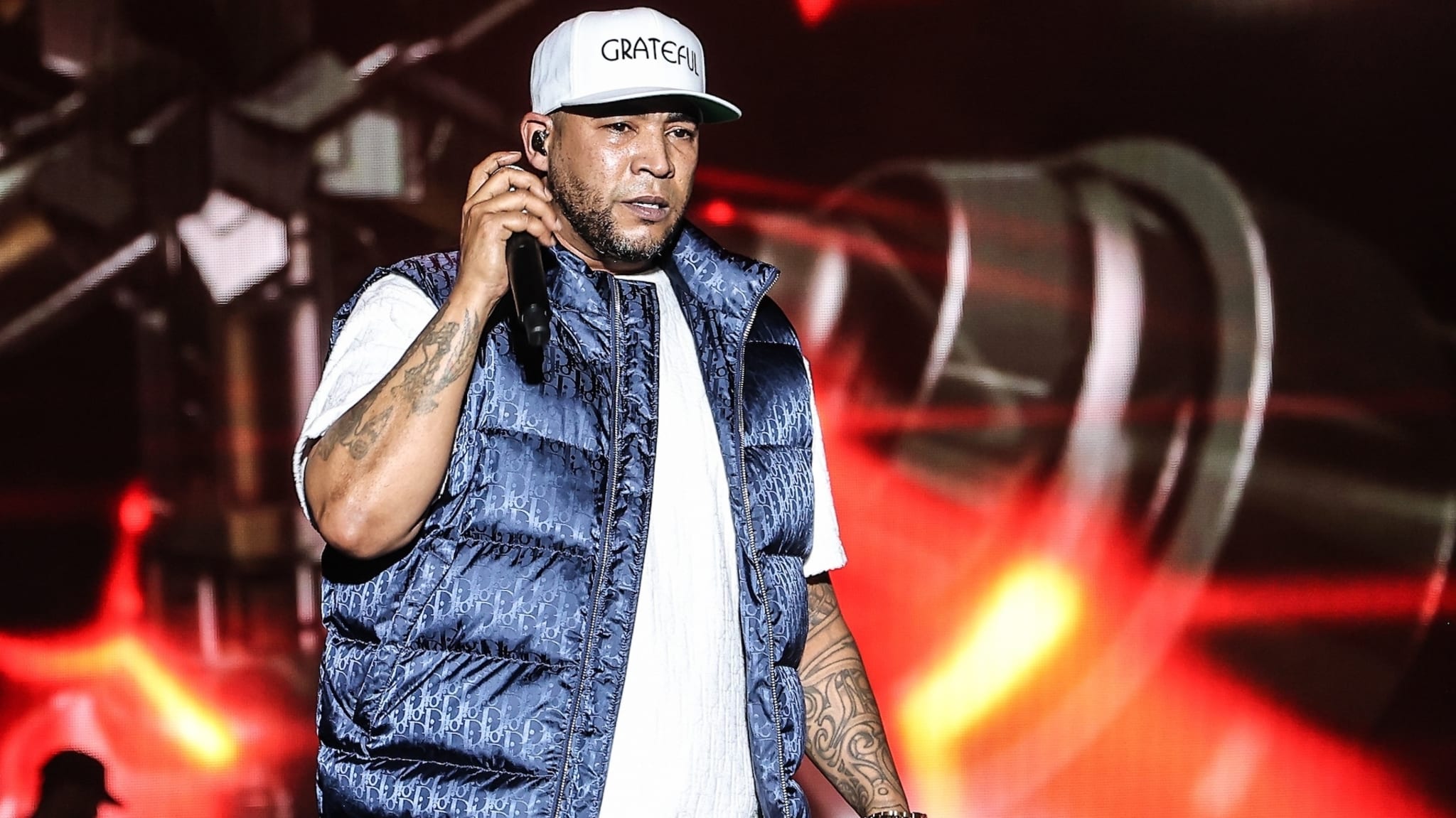 Man wearing a white cap with "GRATEFUL", holding a microphone, dressed in a white shirt and blue puffer vest, performing on stage