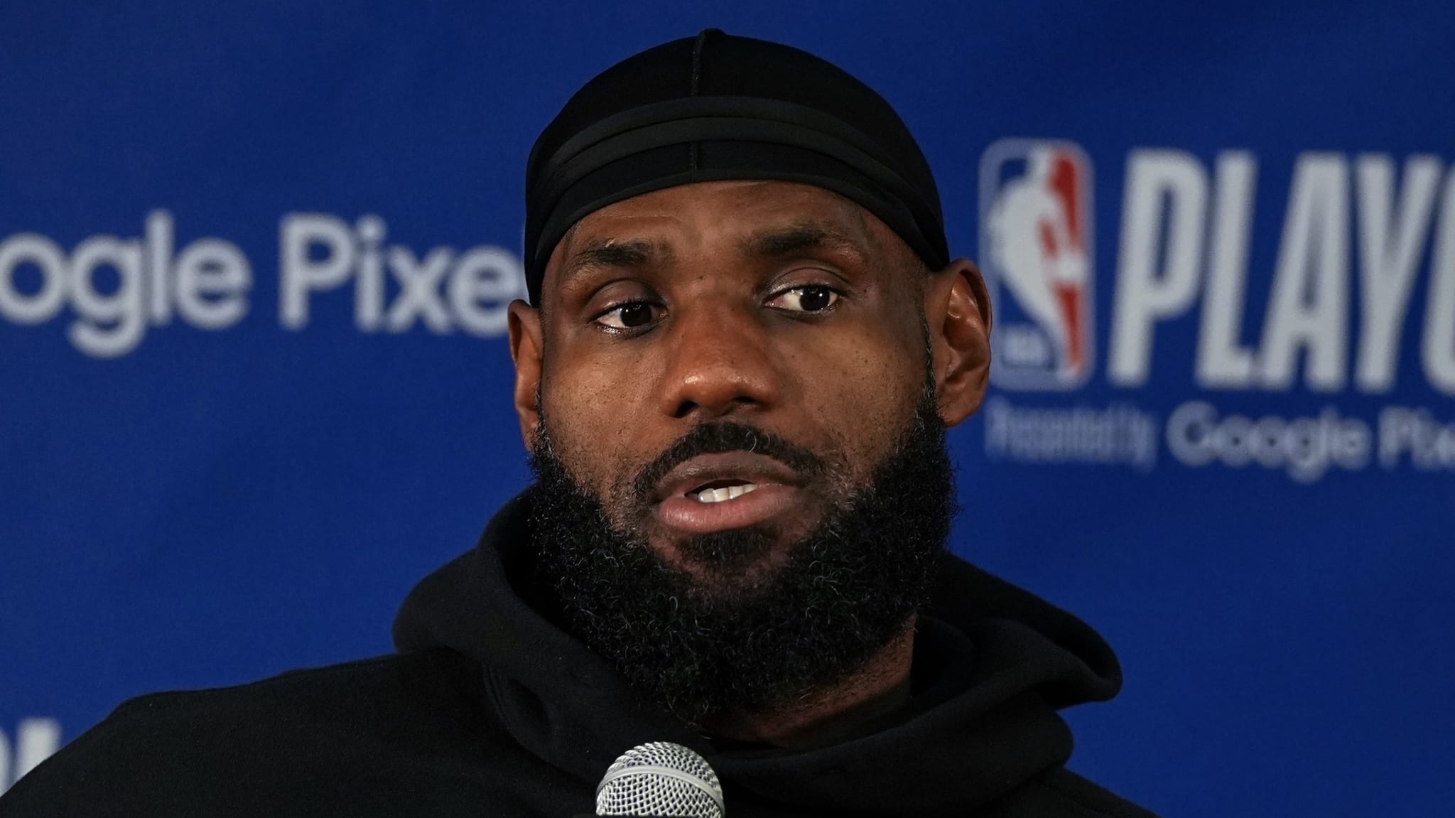 Man in black attire and headband speaks at a press conference with logos in the background