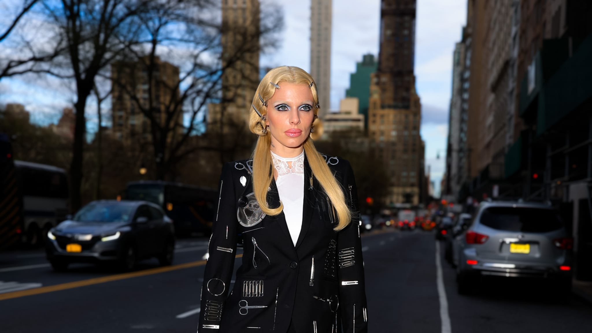 A person with long blonde hair and stylish black clothing stands on a city street with tall buildings and cars in the background